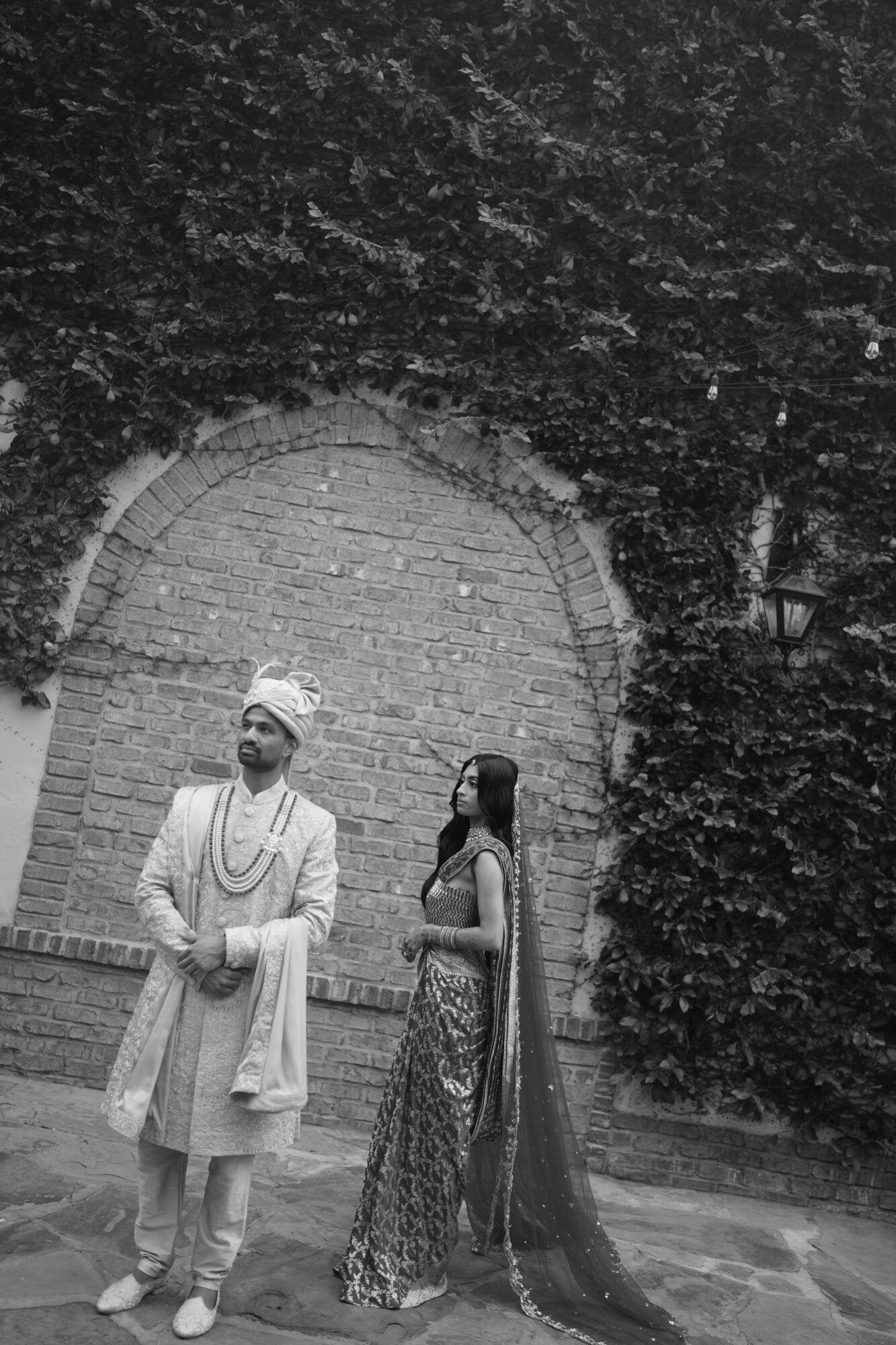 Two people in traditional attire stand outdoors near a brick archway and greenery.