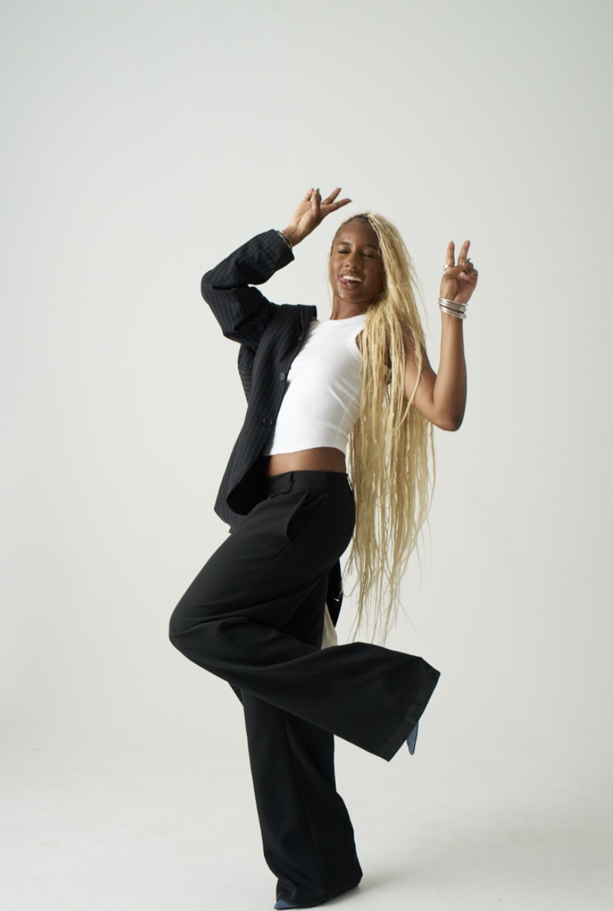 Woman with long hair in a white crop top and black pants poses with one leg raised and arms raised.