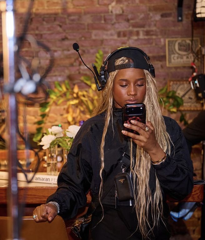 Young woman with long hair wearing headphones, taking a selfie in a brick-walled room with plants and flowers.