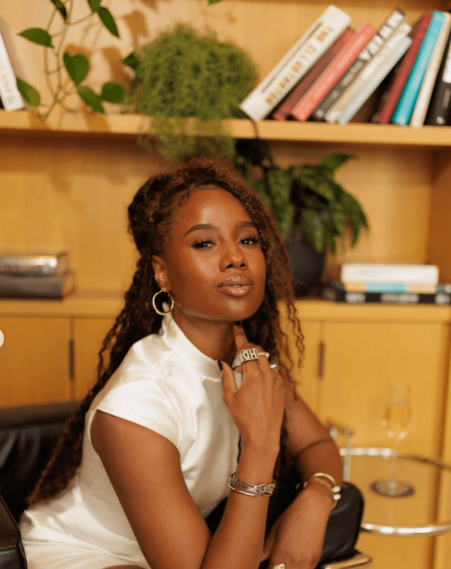 Woman with long curly hair wearing a white top, sitting in a room with books and plants, looking at the camera.