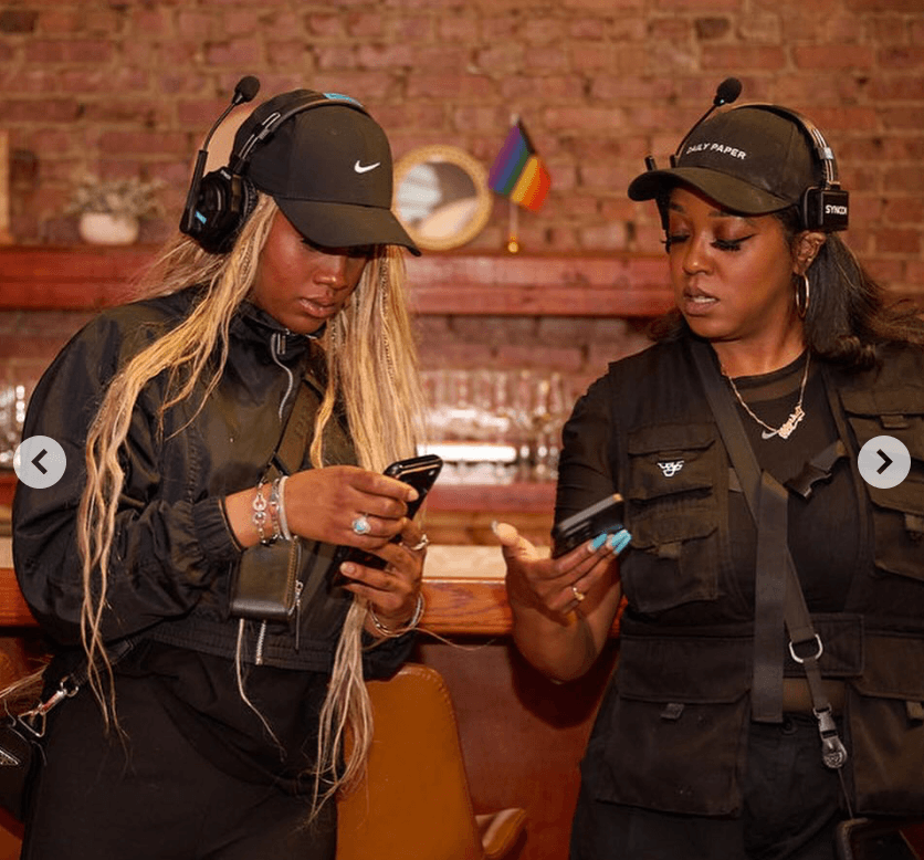 Two women wearing headphones and caps look at their phones in a brick-walled setting with a rainbow flag in the background.