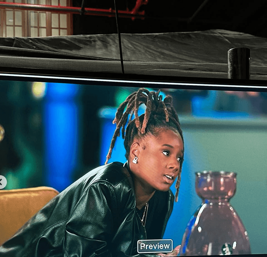 Woman with dreadlocks wearing a leather jacket, sitting indoors with a blurred background and a purple vase.