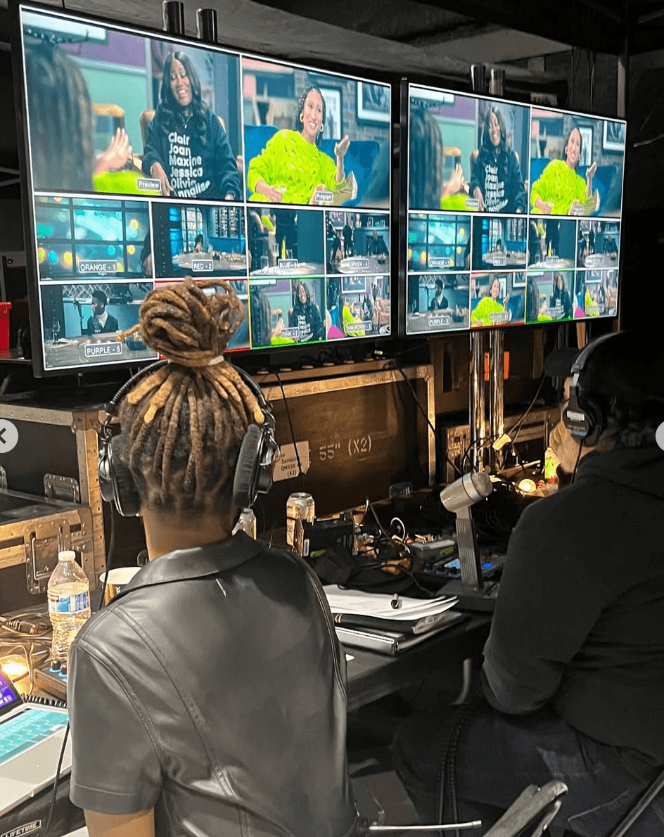 Two people with headphones sit at a table with multiple monitors displaying a woman in a bright green shirt on a video call.