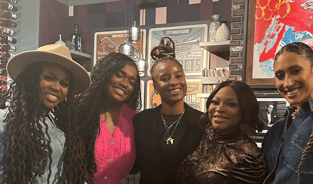 Five women smiling together indoors, background includes books and artwork, casual attire, diverse hairstyles, and accessories.