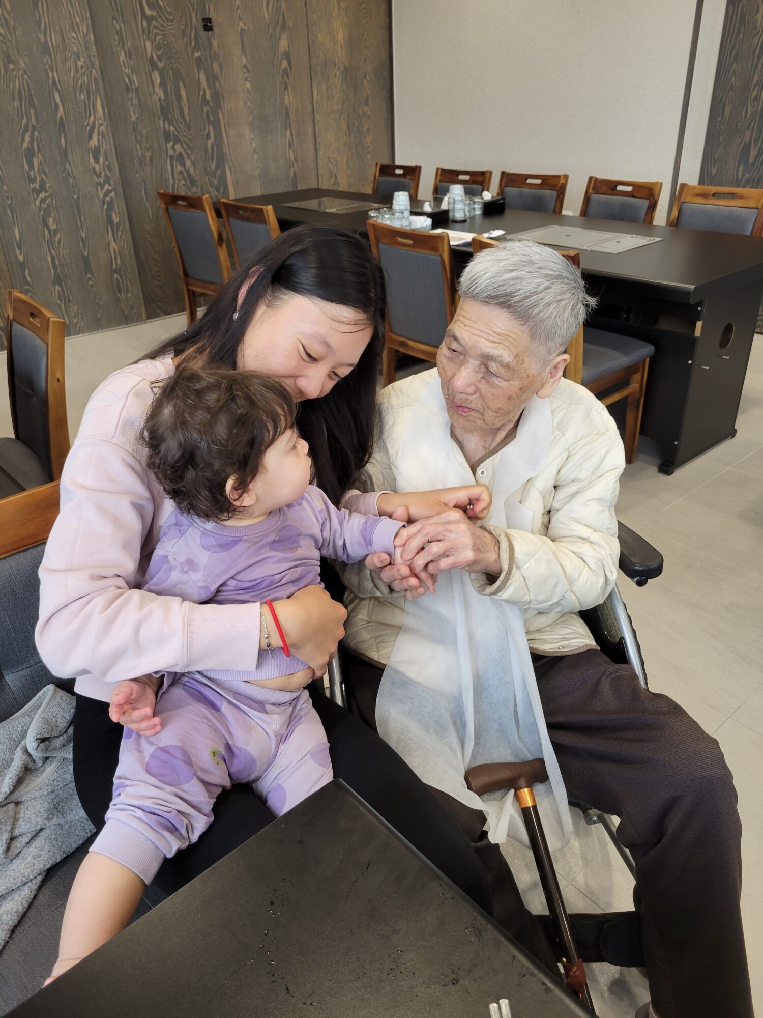 An elderly woman in a wheelchair holding hands with a young girl, with another girl leaning on the table, in a room with wooden chairs and tables.