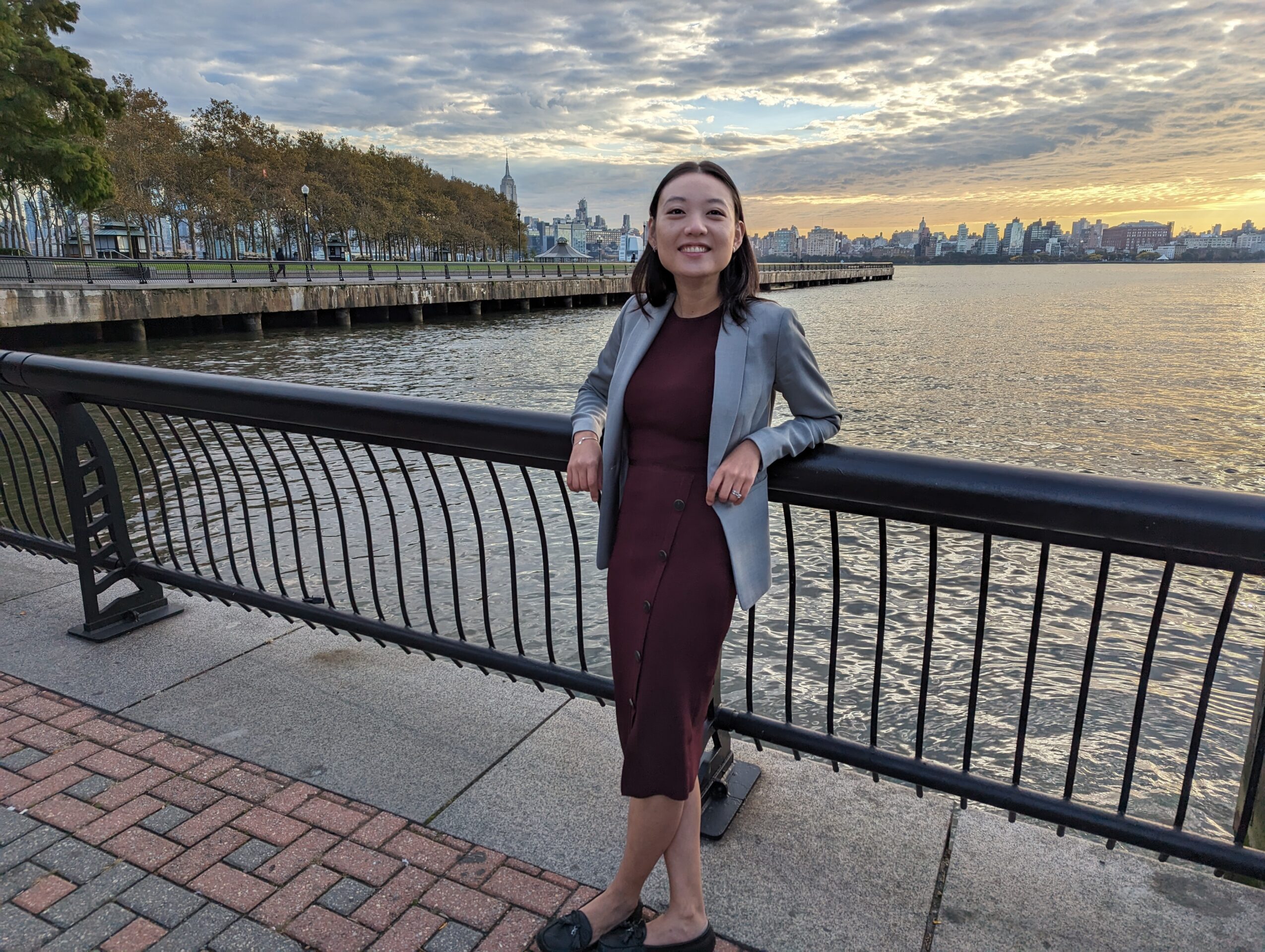 Woman standing by a railing near water with city skyline and cloudy sky in background, smiling, wearing a dress and blazer.