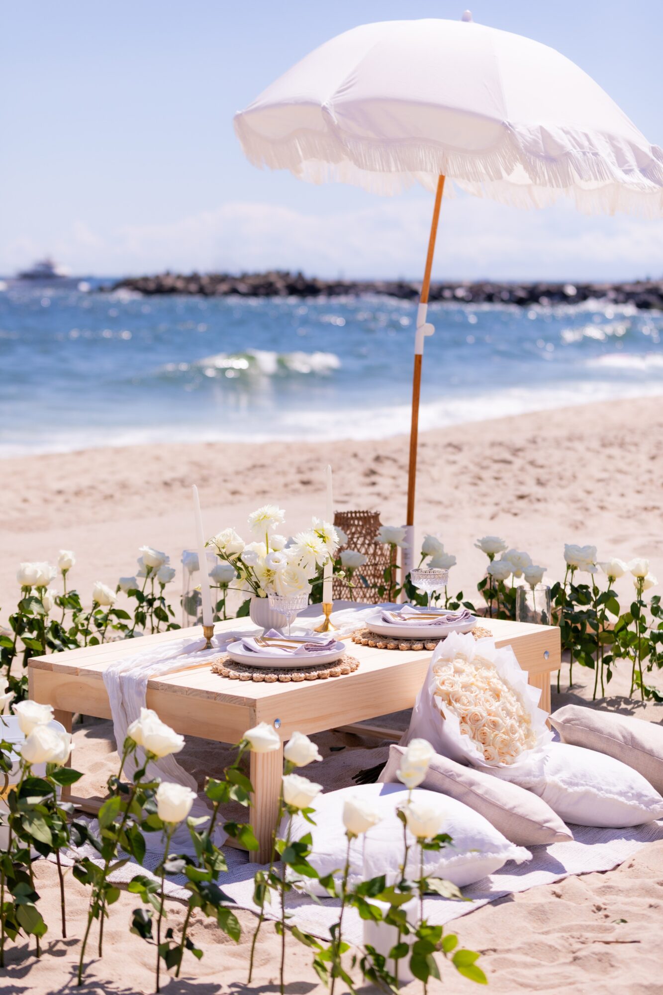 Beachside table with white flowers, cushions, and a parasol on sandy beach, ocean in background, sunny day.