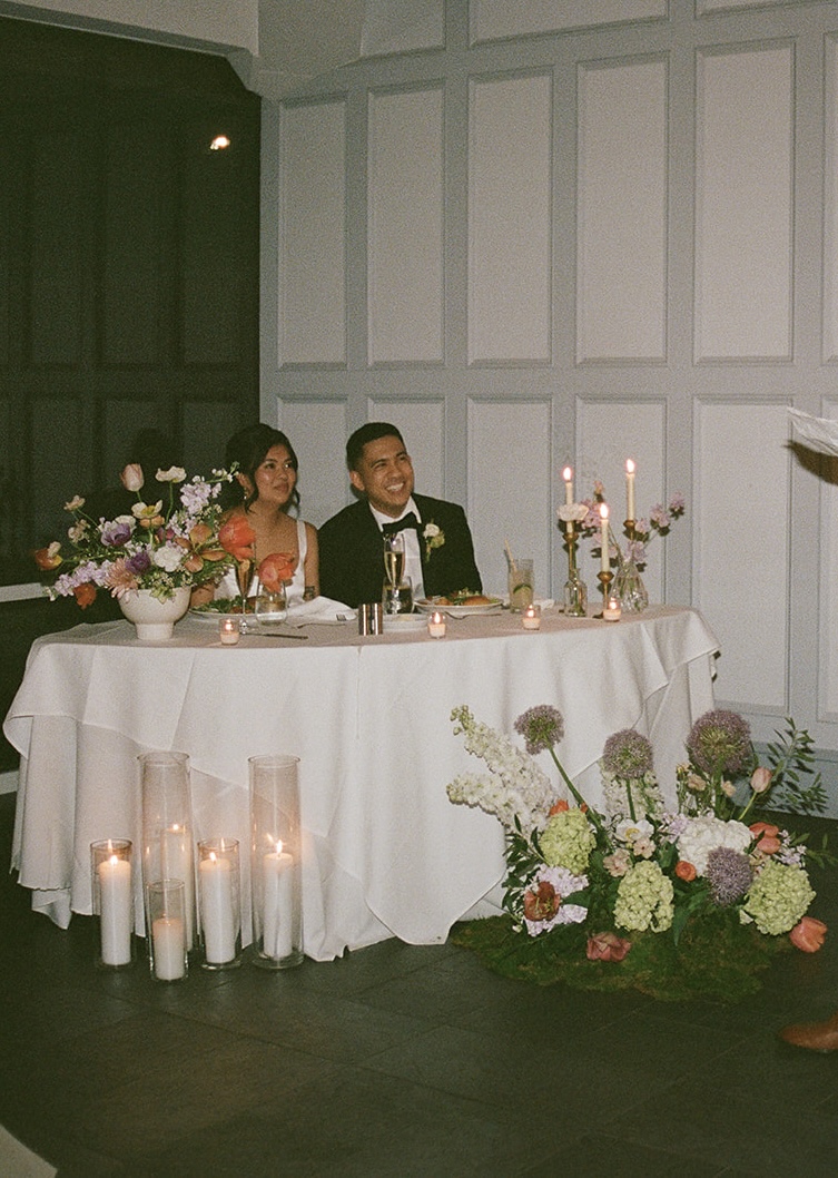 Two people sit at a decorated table with candles and flowers, in a room with paneled walls, during a formal event.
