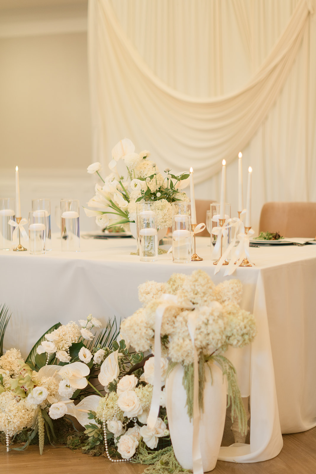 Elegant table with white floral centerpiece and candles, decorated with white drapery in the background.