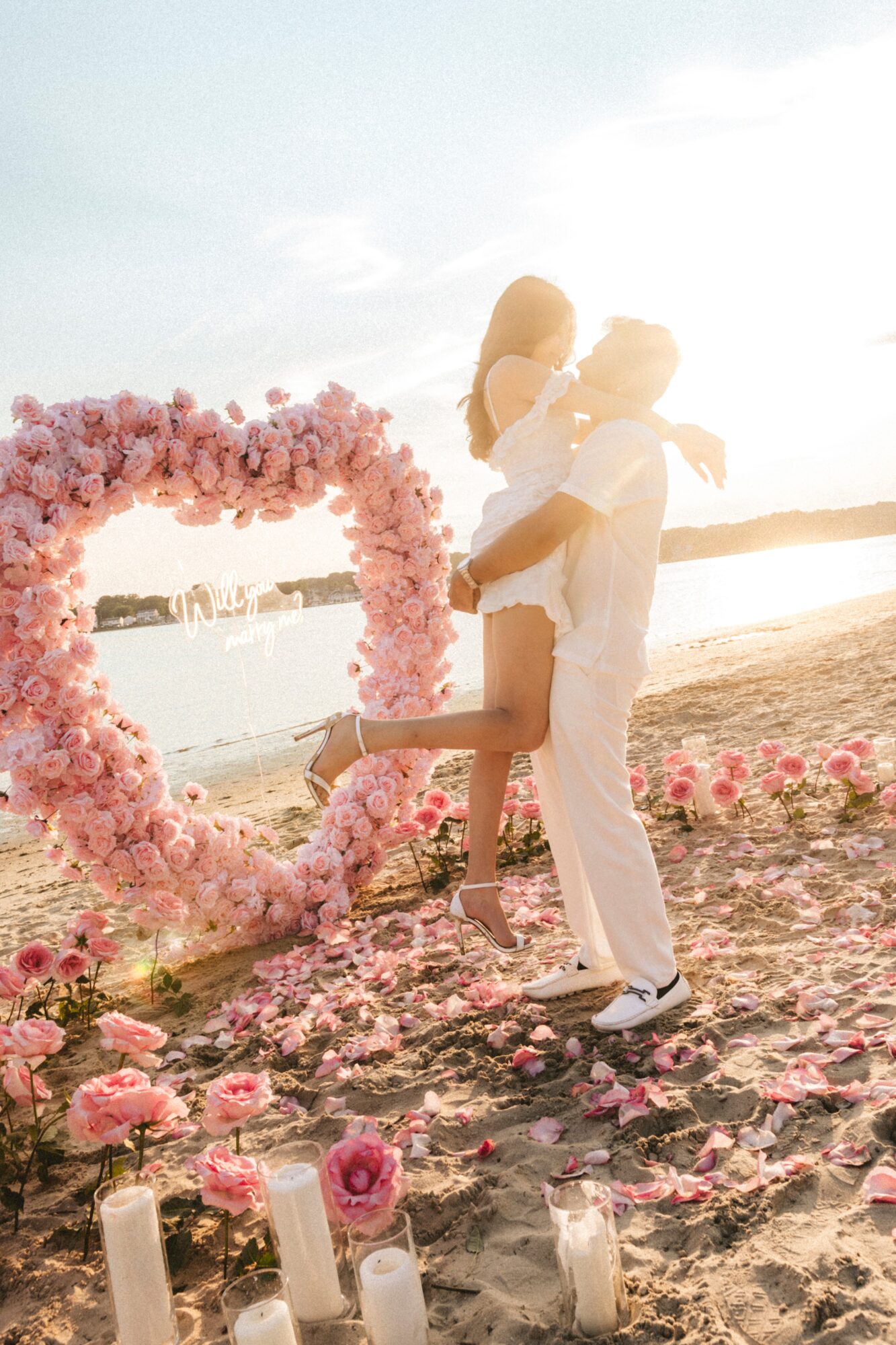 Couple dancing on beach near heart-shaped floral arch, with candles and flowers on sand, during sunset.