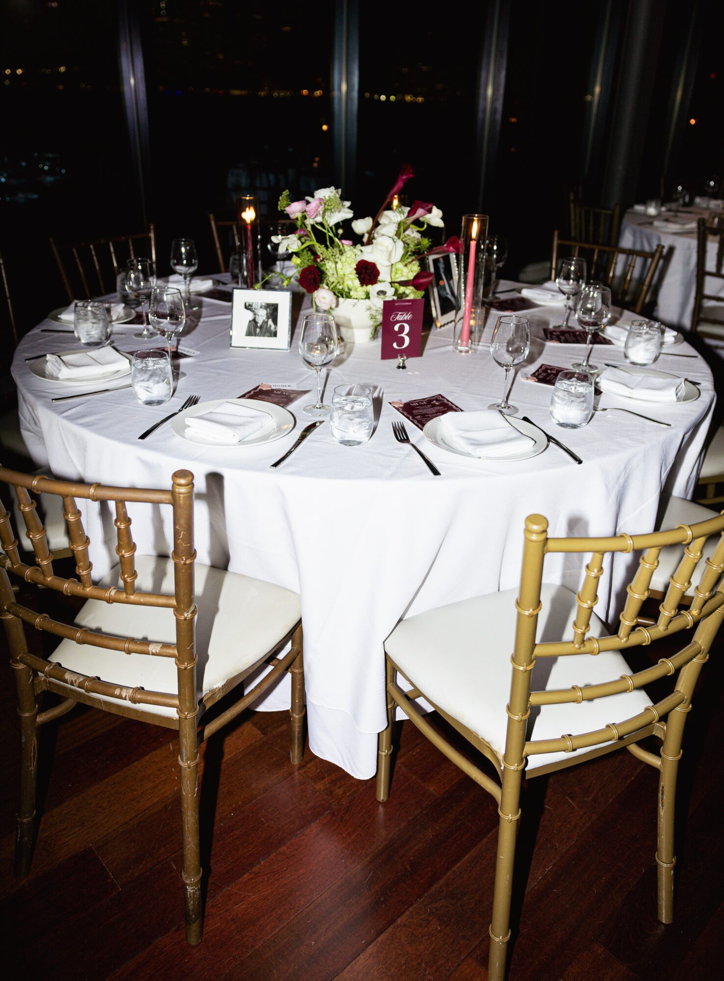 Round table with white tablecloth, floral centerpiece, place settings, and chairs in a dimly lit room with large windows.