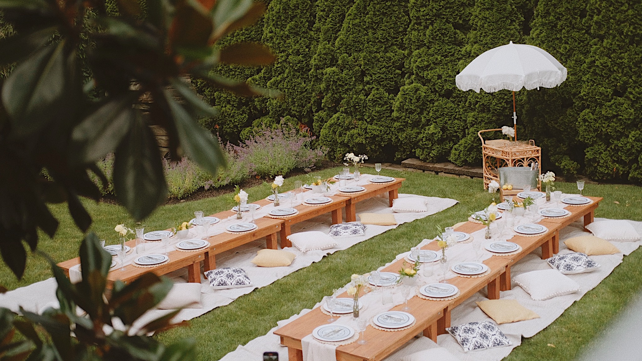 Outdoor dining setup with long wooden tables, white plates, and cushions on grass, with a white umbrella and green hedge background.
