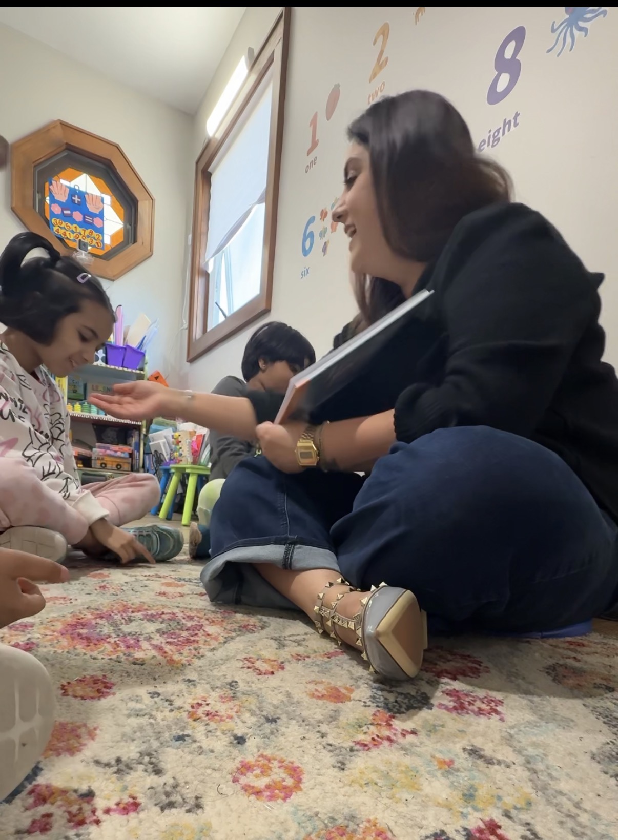 Woman sitting on the floor with children, engaging in a classroom activity, with colorful numbers on the wall and a window.