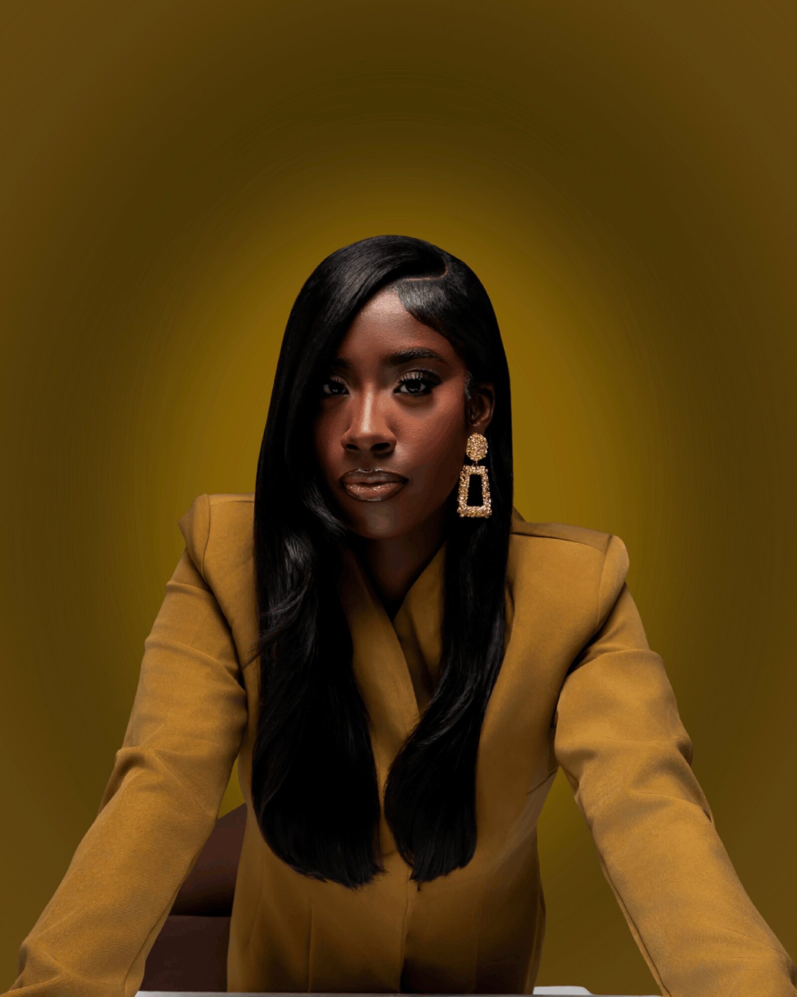 A woman with long black hair wearing a mustard-colored blazer and large earrings, posed against a yellow background.