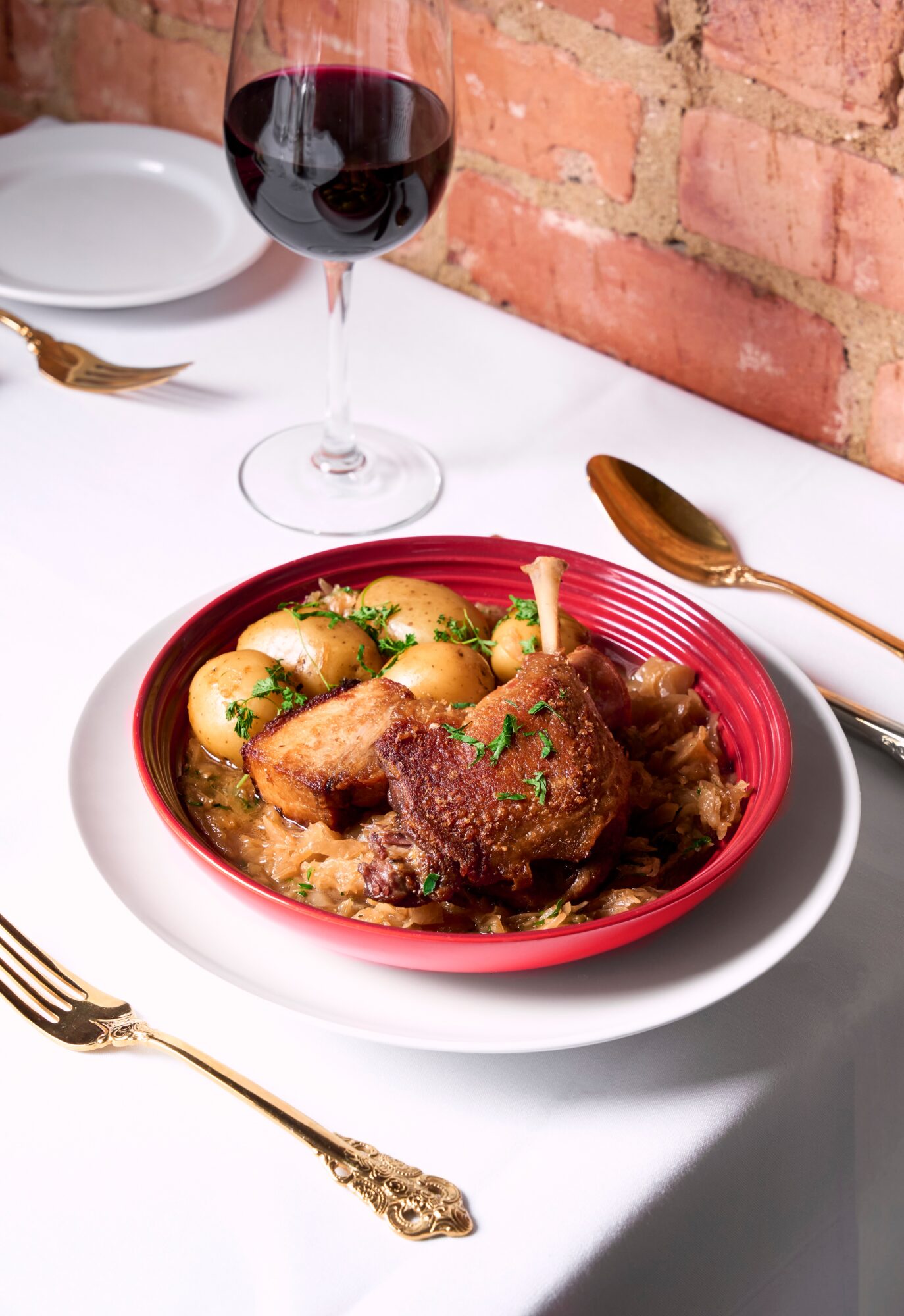 Plate of cooked meat and potatoes with a glass of red wine on a white table against a brick wall.