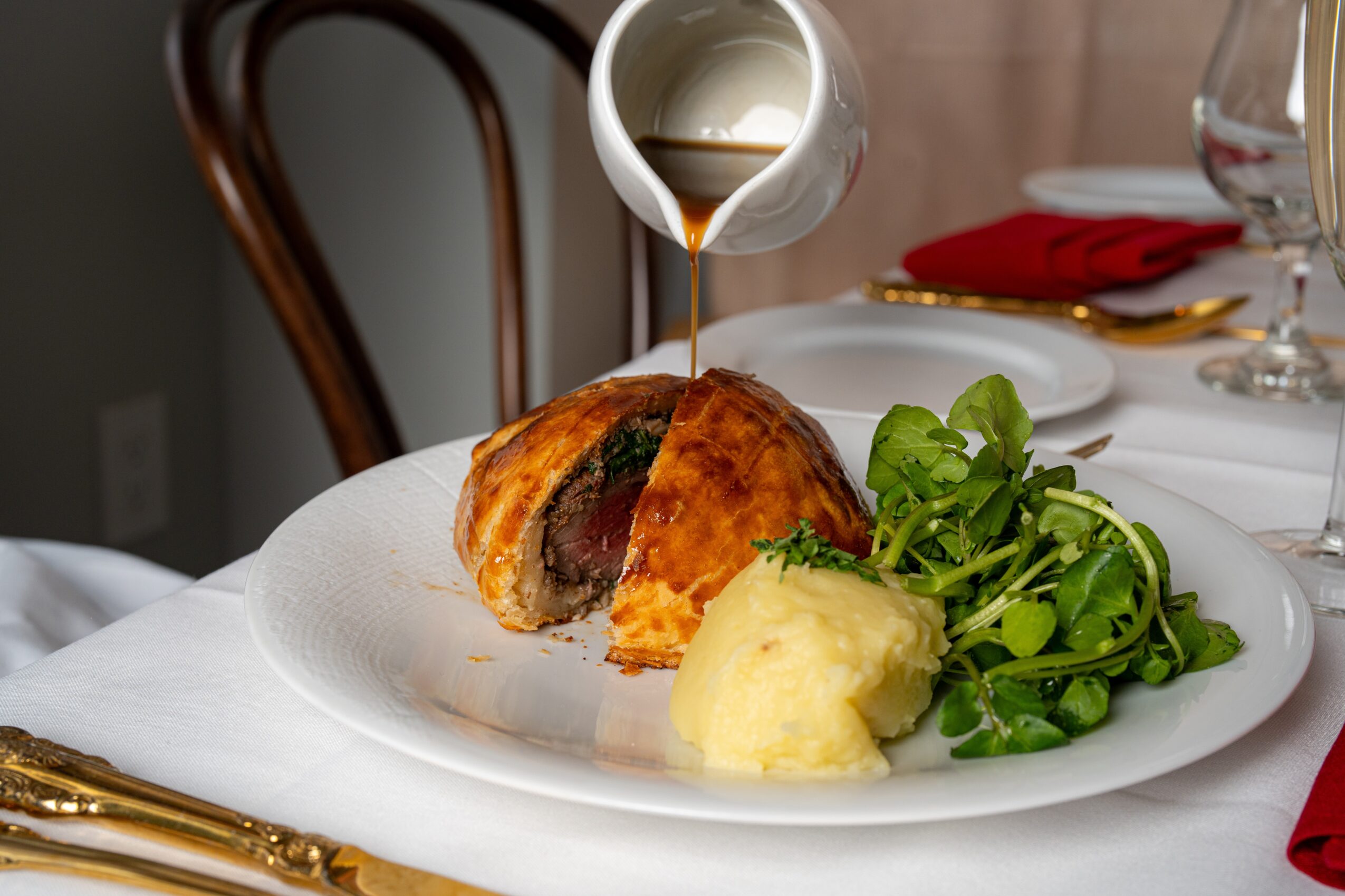 A plated beef Wellington with mashed potatoes and greens, with gravy being poured over the dish, on a white tablecloth.