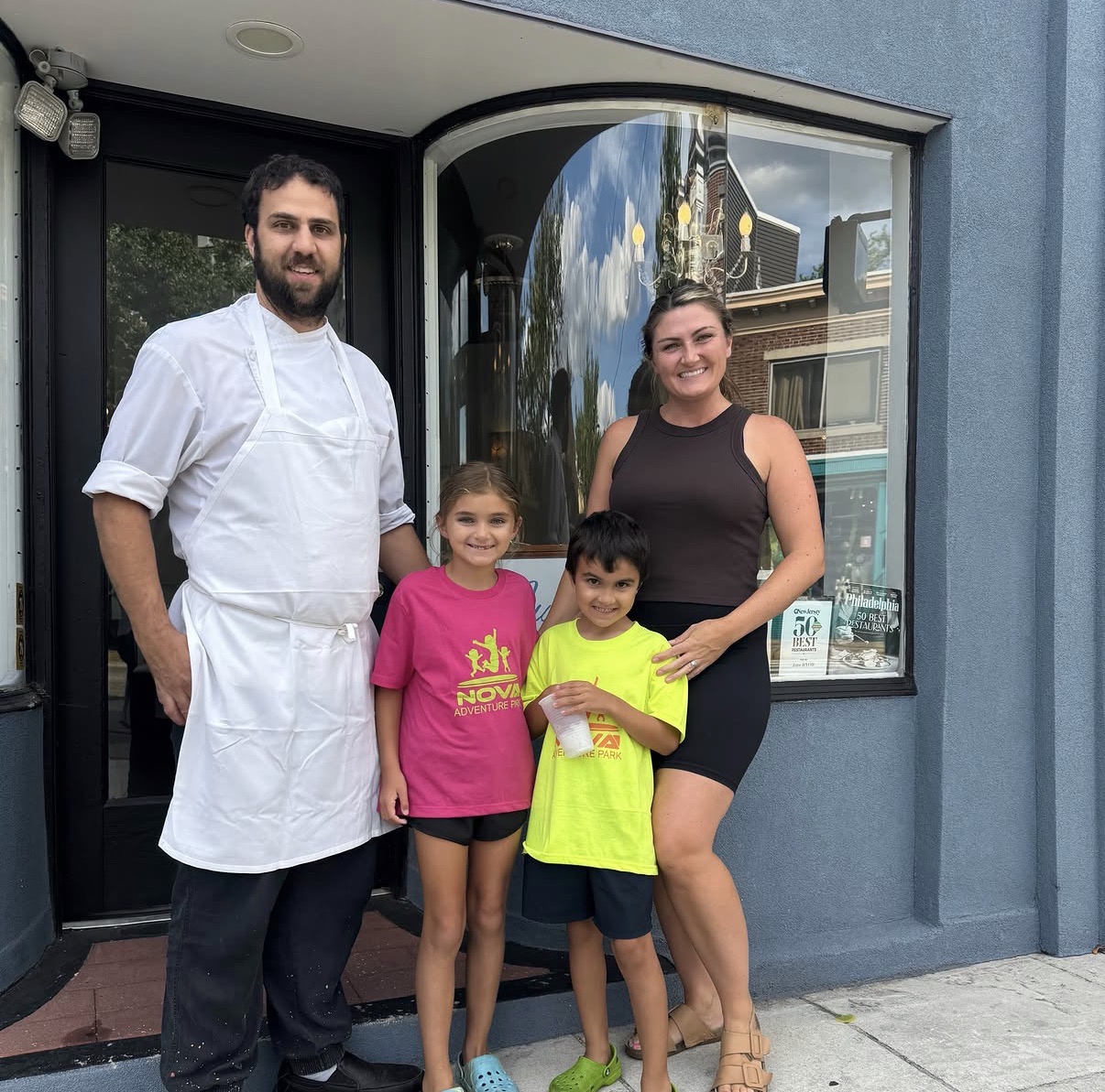 Four people, two adults and two children, stand outside a building with a large window, smiling at the camera.