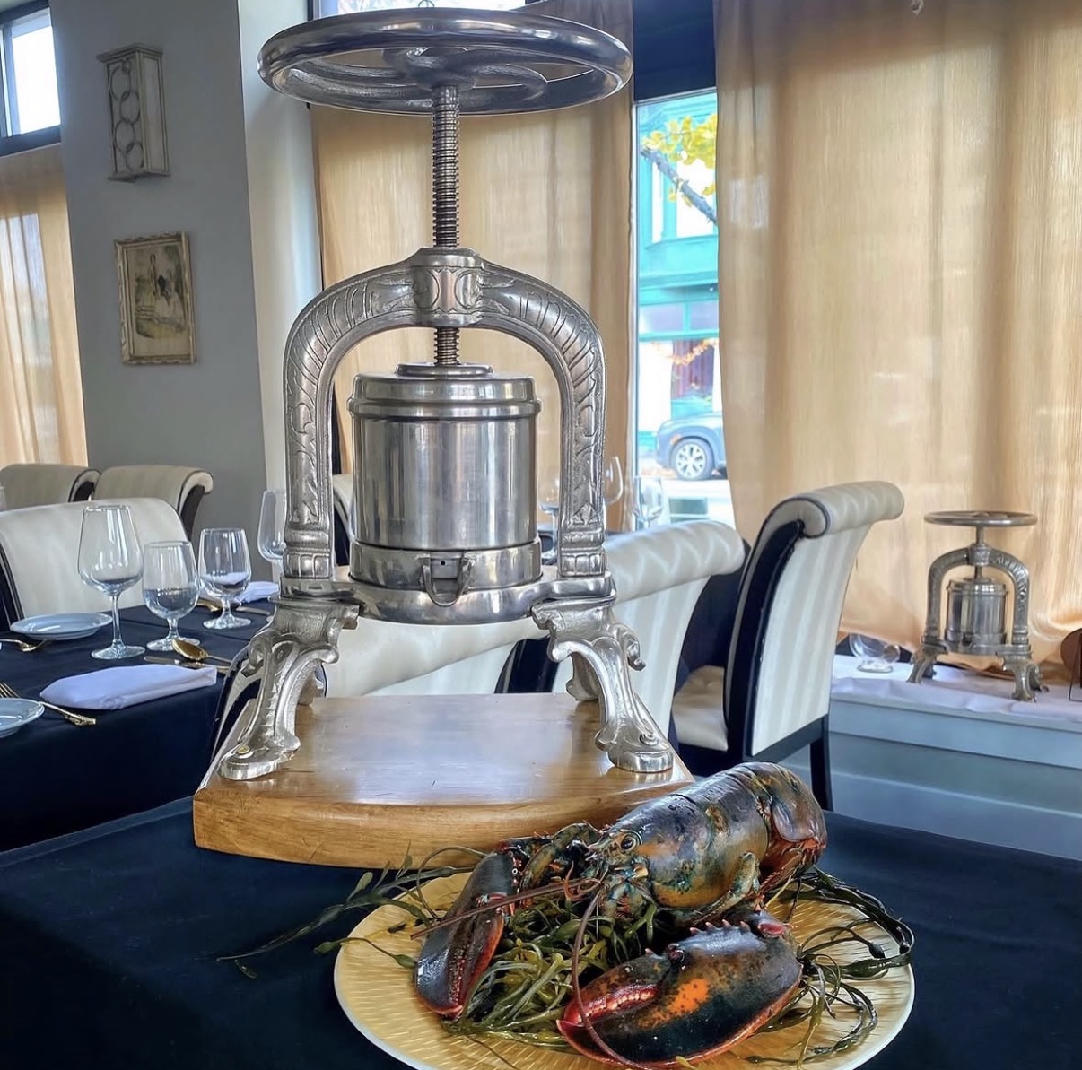A large metal lobster cracker on a wooden table with a plate of cooked lobster and seafood, in a dining room with curtains and chairs.