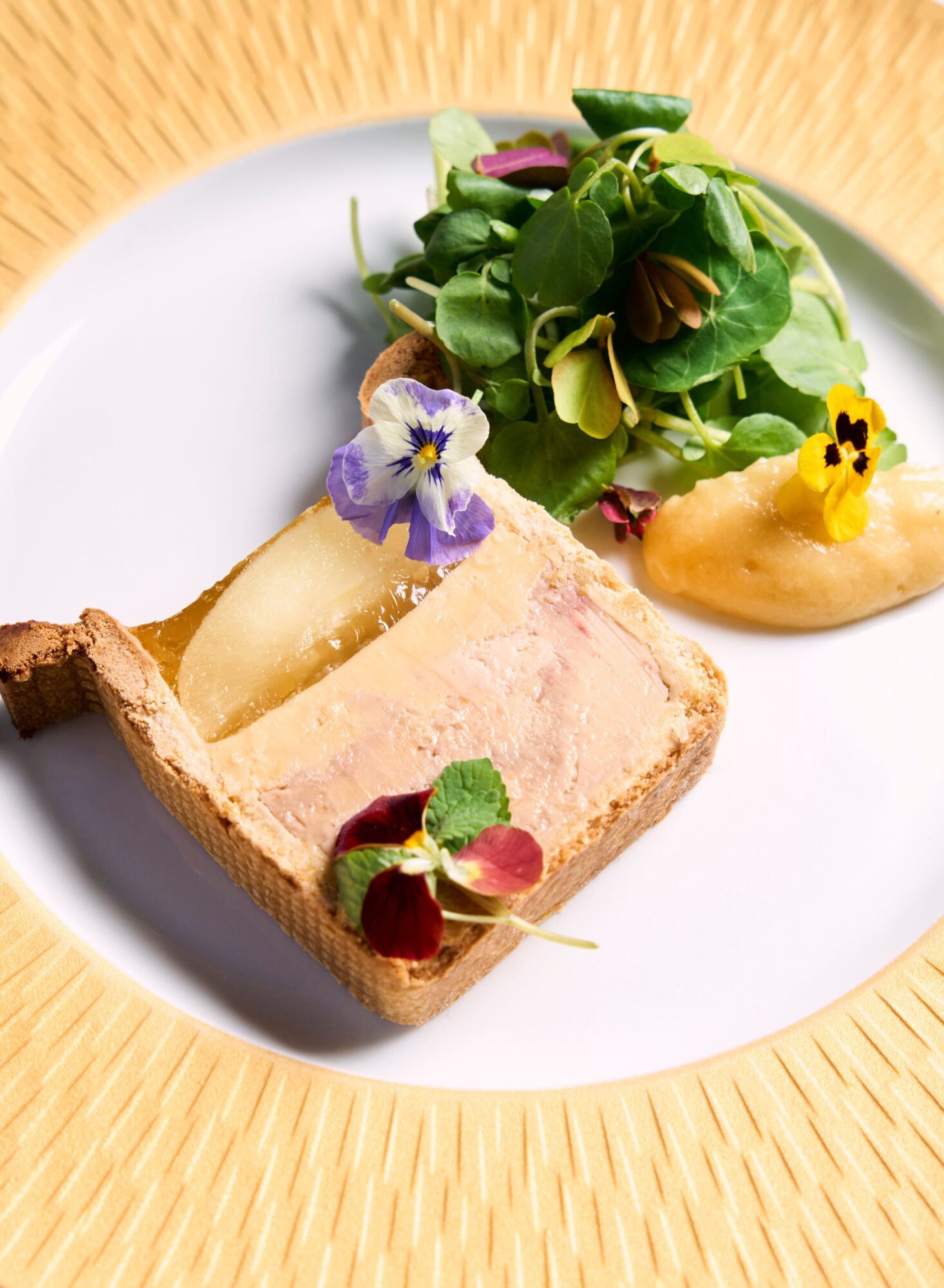 A slice of pâté with a side salad and edible flowers on a white plate, on a woven placemat.