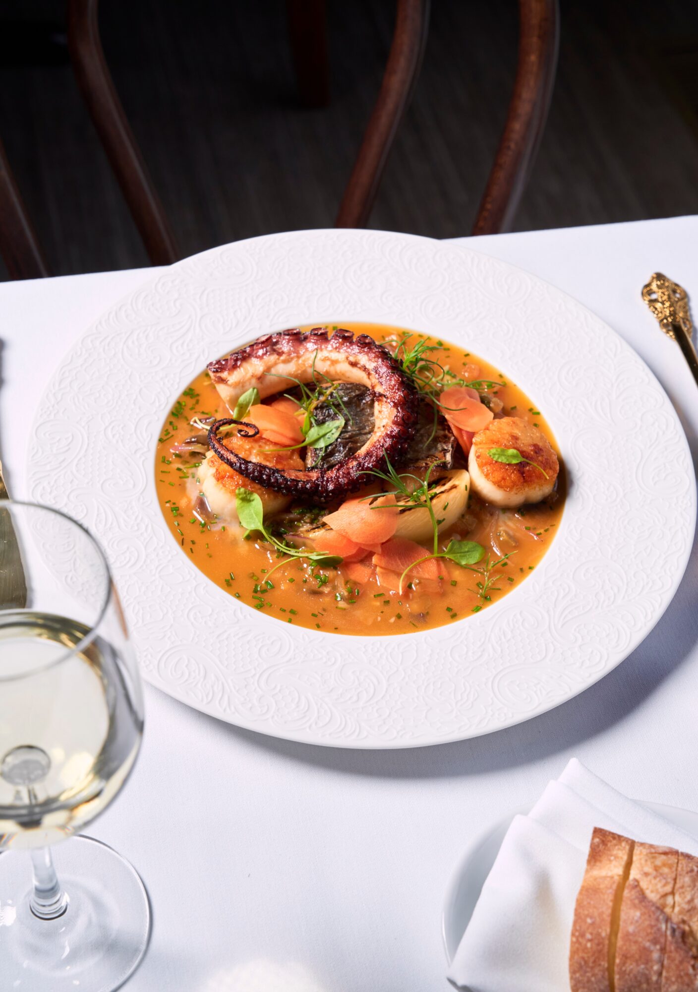 Seafood dish with octopus, shrimp, and vegetables in broth on a white plate, set on a white tablecloth.