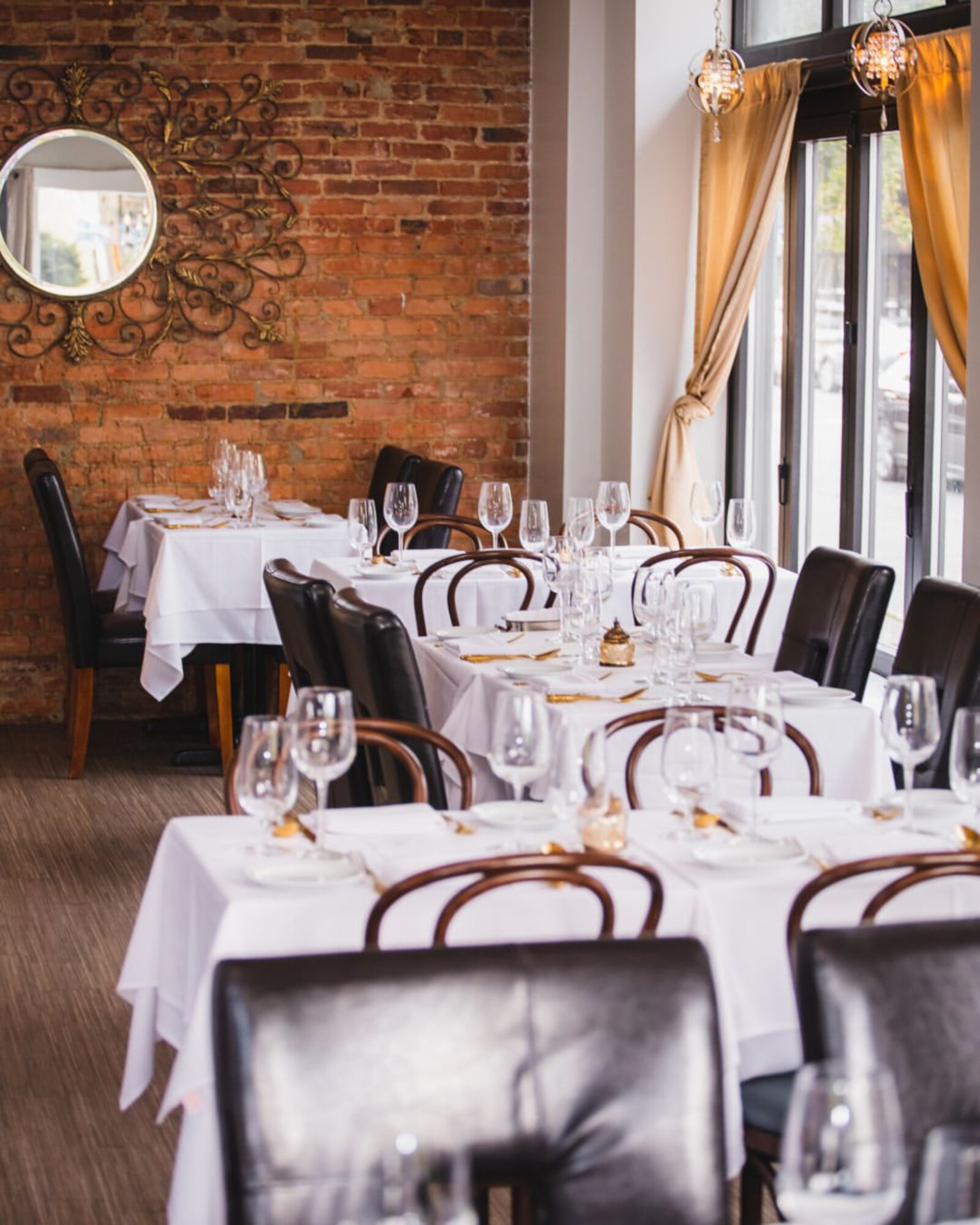 Elegant restaurant interior with white tablecloths, glassware, and black chairs, brick wall, and large windows with yellow curtains.