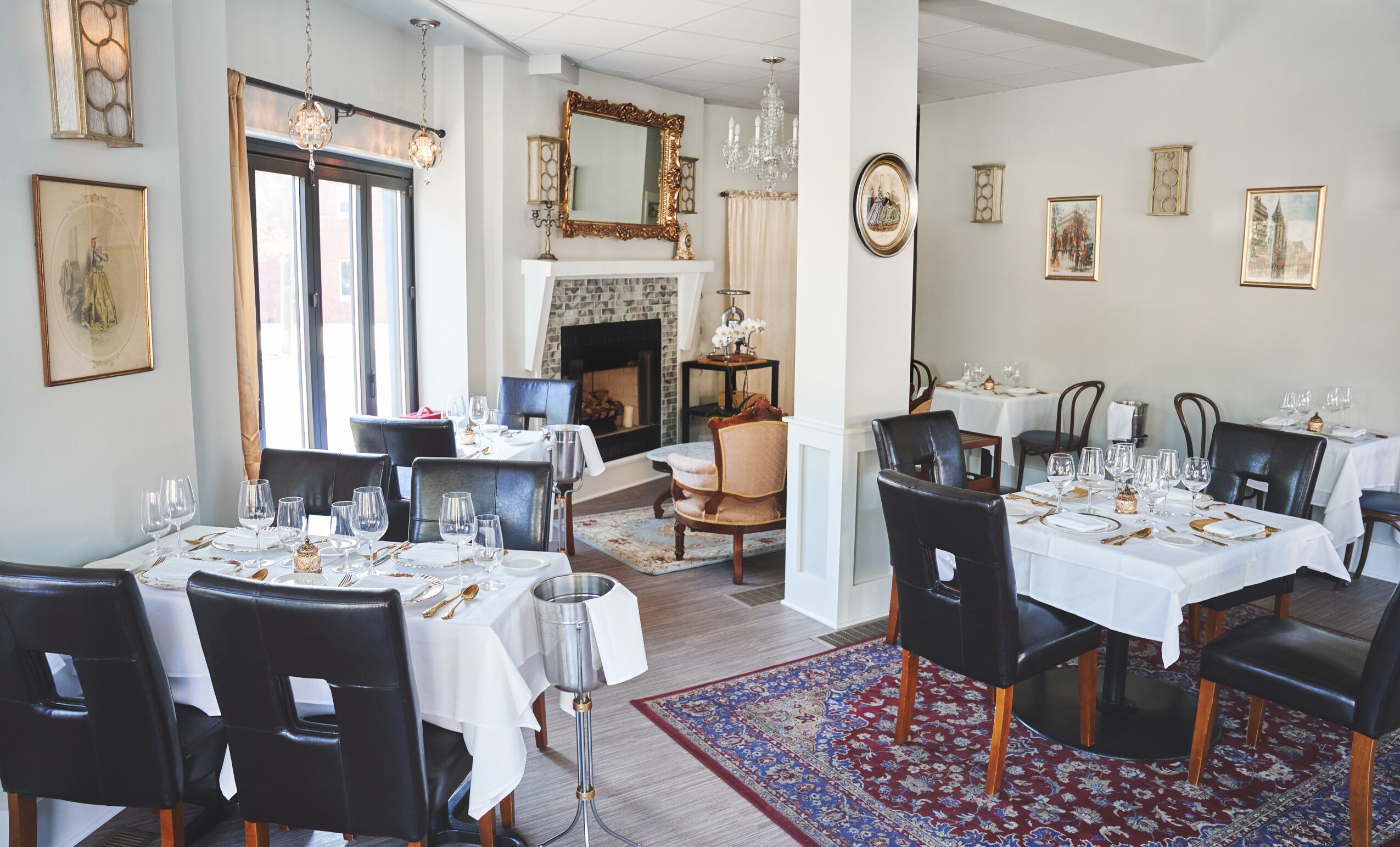 Elegant dining room with white tablecloths, black chairs, and wall art, featuring a fireplace and large windows.