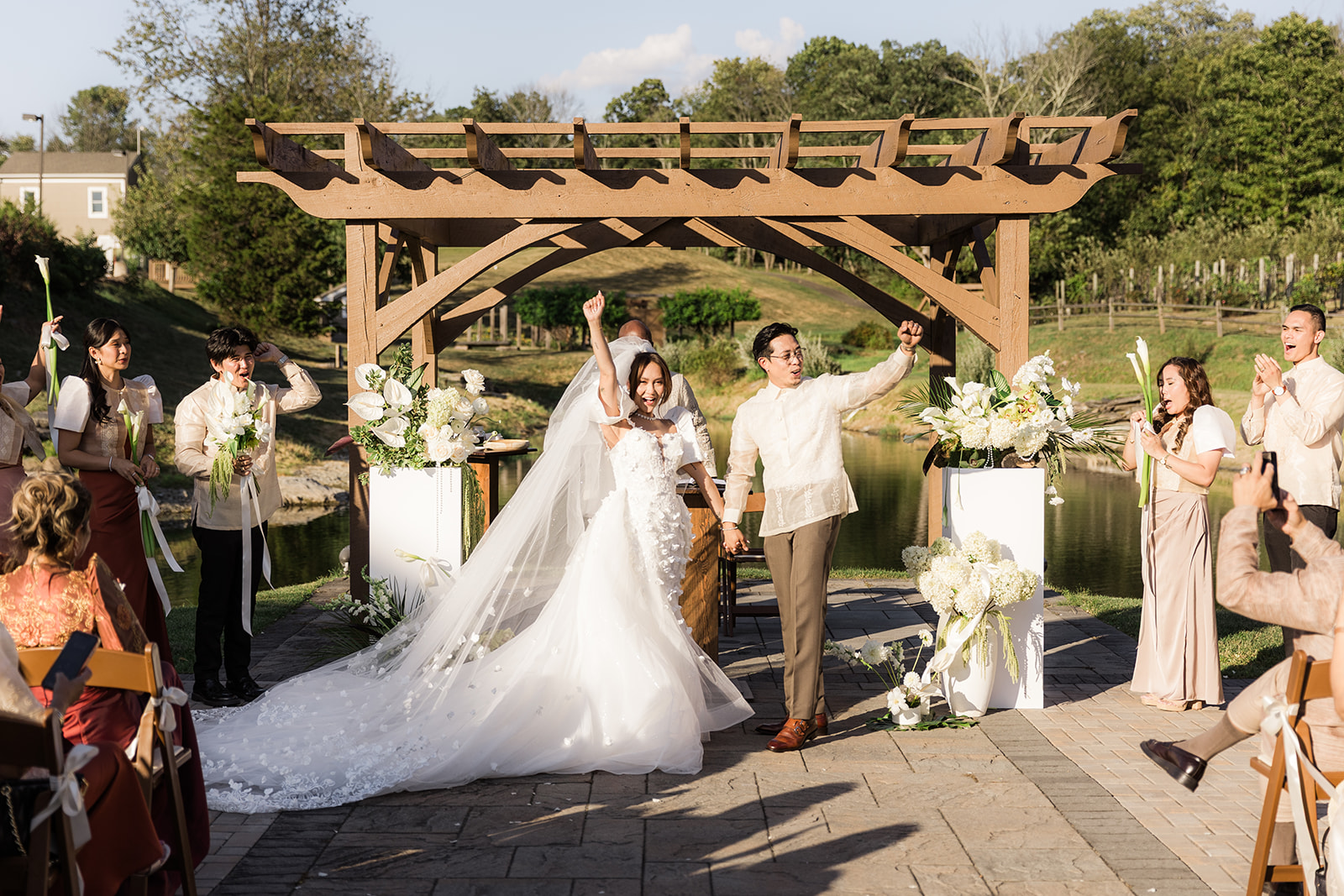 Bride and groom holding hands under a wooden arch at an outdoor wedding ceremony, guests seated nearby, trees in background.