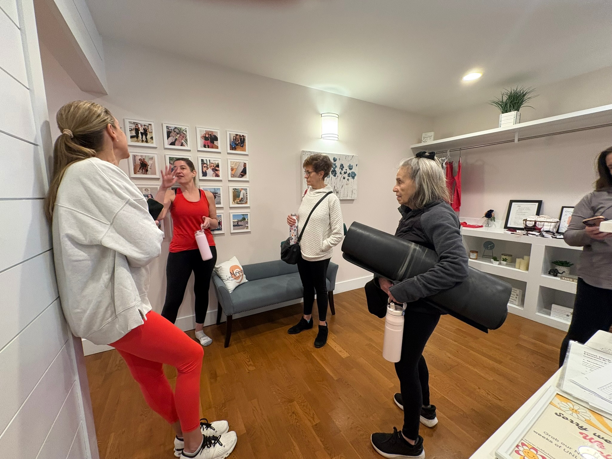 Group of five women in a room with photos on the wall, some holding items, engaged in conversation.