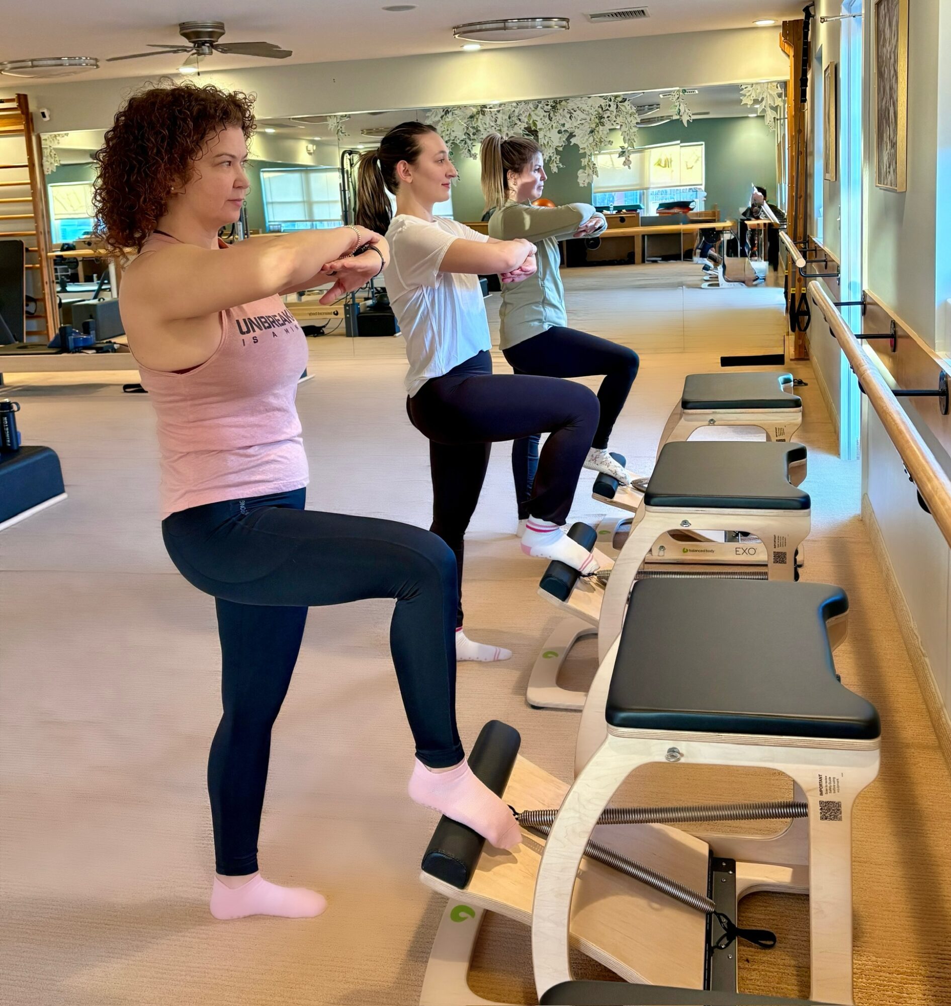 Three women in a fitness class using exercise machines, standing on one leg, in a bright studio with mirrors and windows.