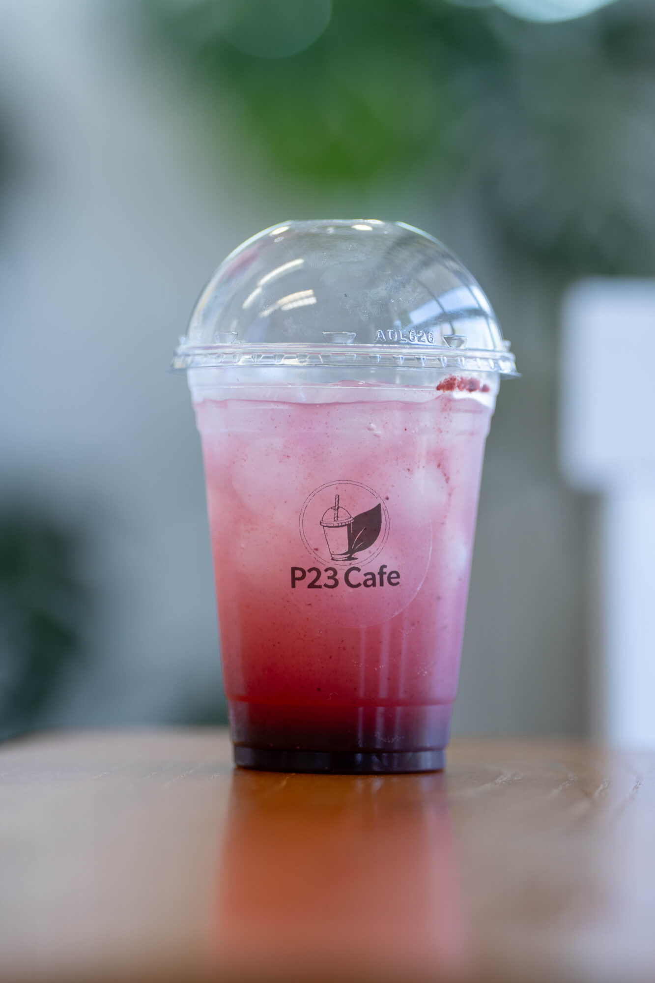 Pink and red layered beverage in a clear plastic cup with a domed lid, on a wooden surface, blurred background.