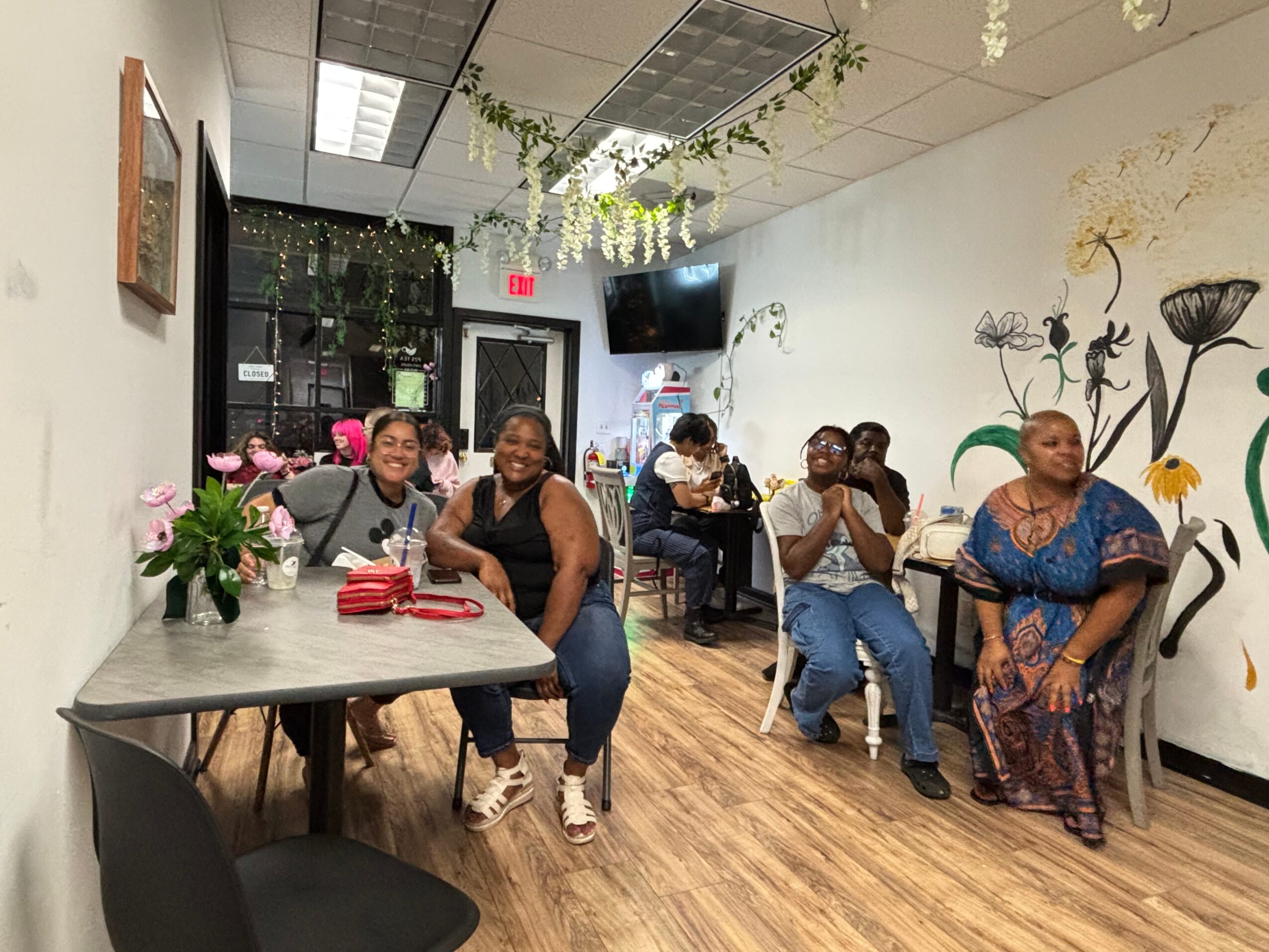 Group of women sitting and smiling in a room with floral wall art and wooden flooring.