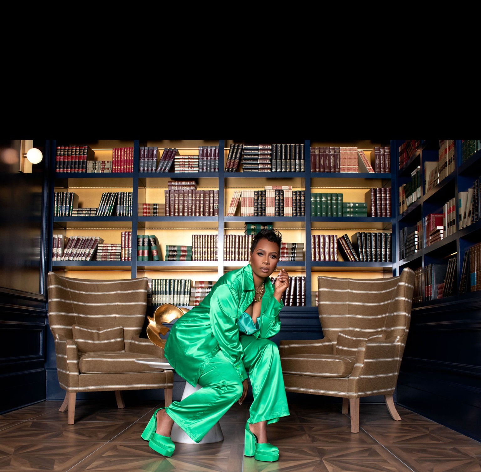 Woman in green outfit and platform shoes crouches in a library with bookshelves and two beige sofas behind her.