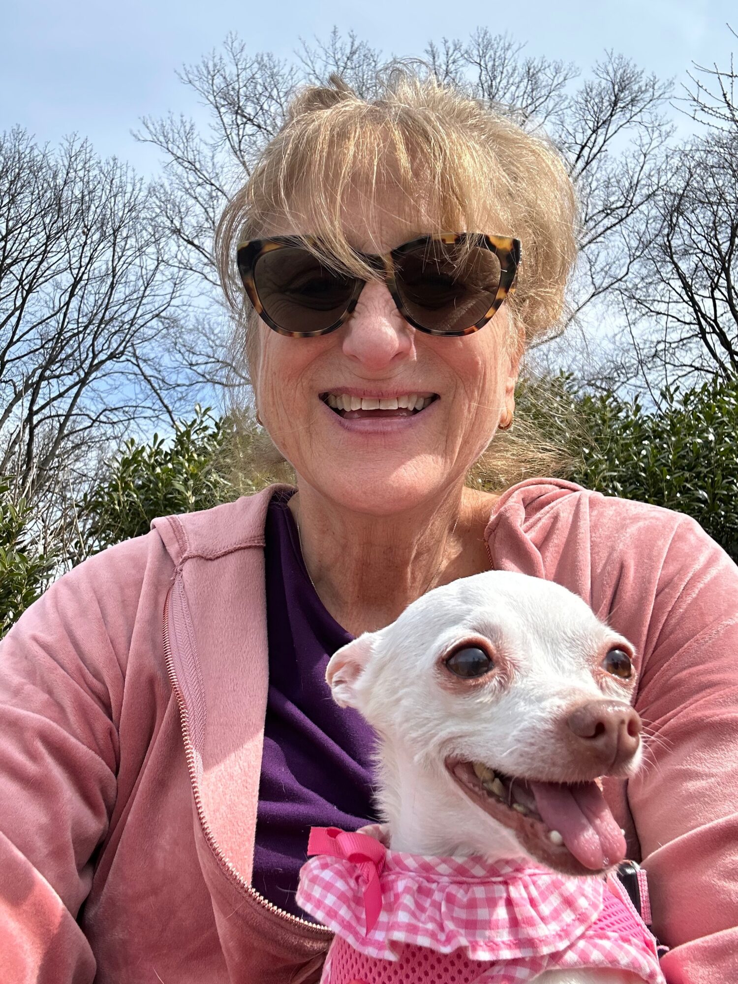 Smiling woman wearing sunglasses outdoors with a small dog wearing a pink ruffled collar, trees in background.