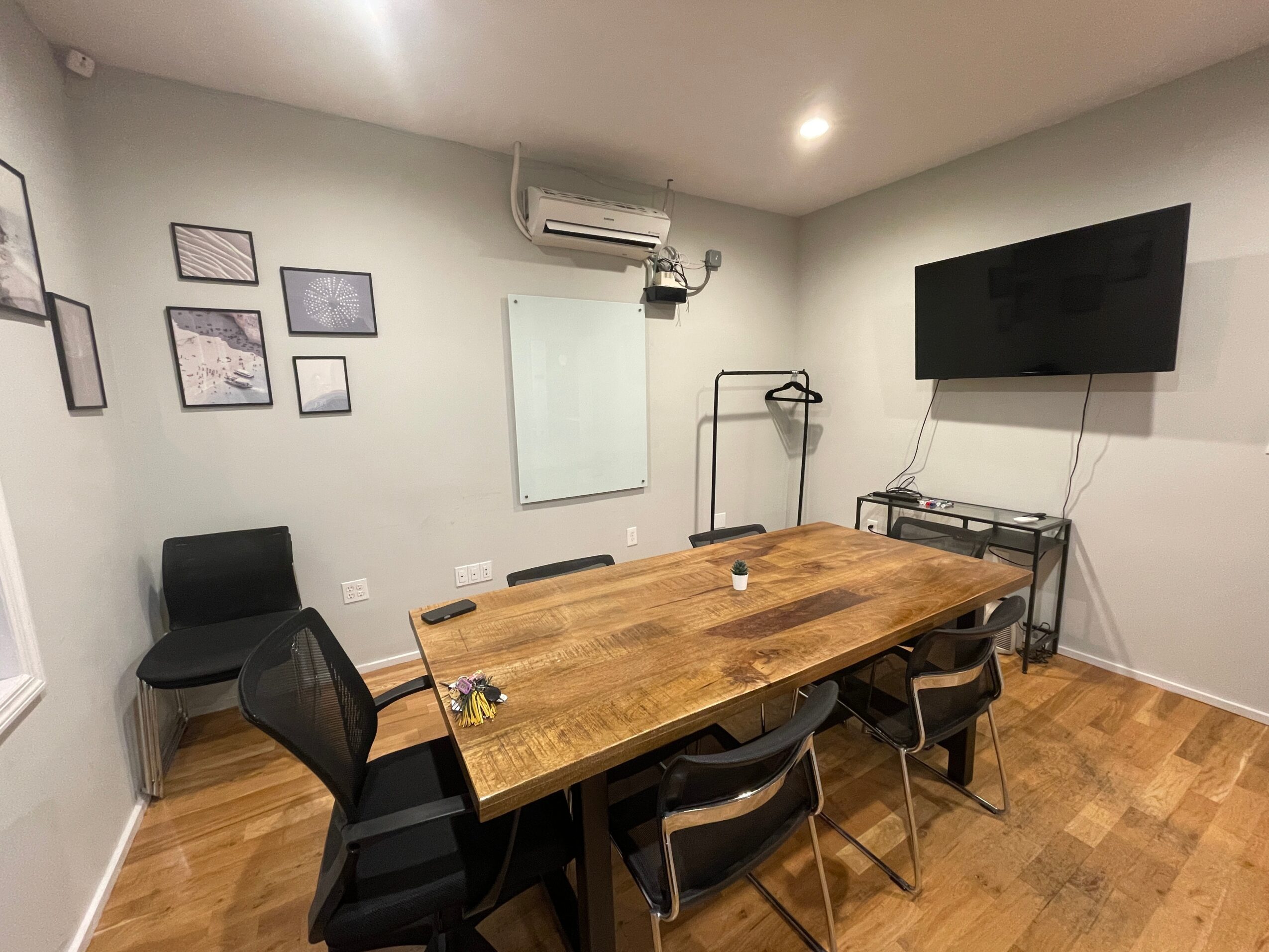 Conference room with wooden table, six chairs, wall-mounted TV, whiteboard, and framed pictures on light-colored walls.