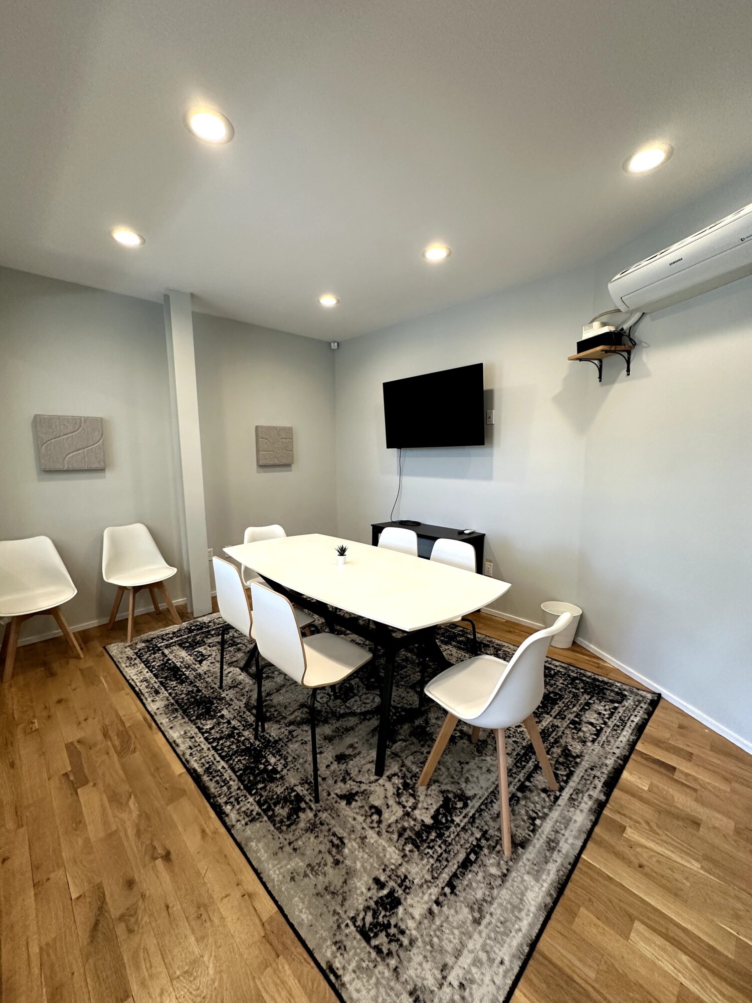 Conference room with a white oval table, six chairs, a wall-mounted TV, and a patterned rug.