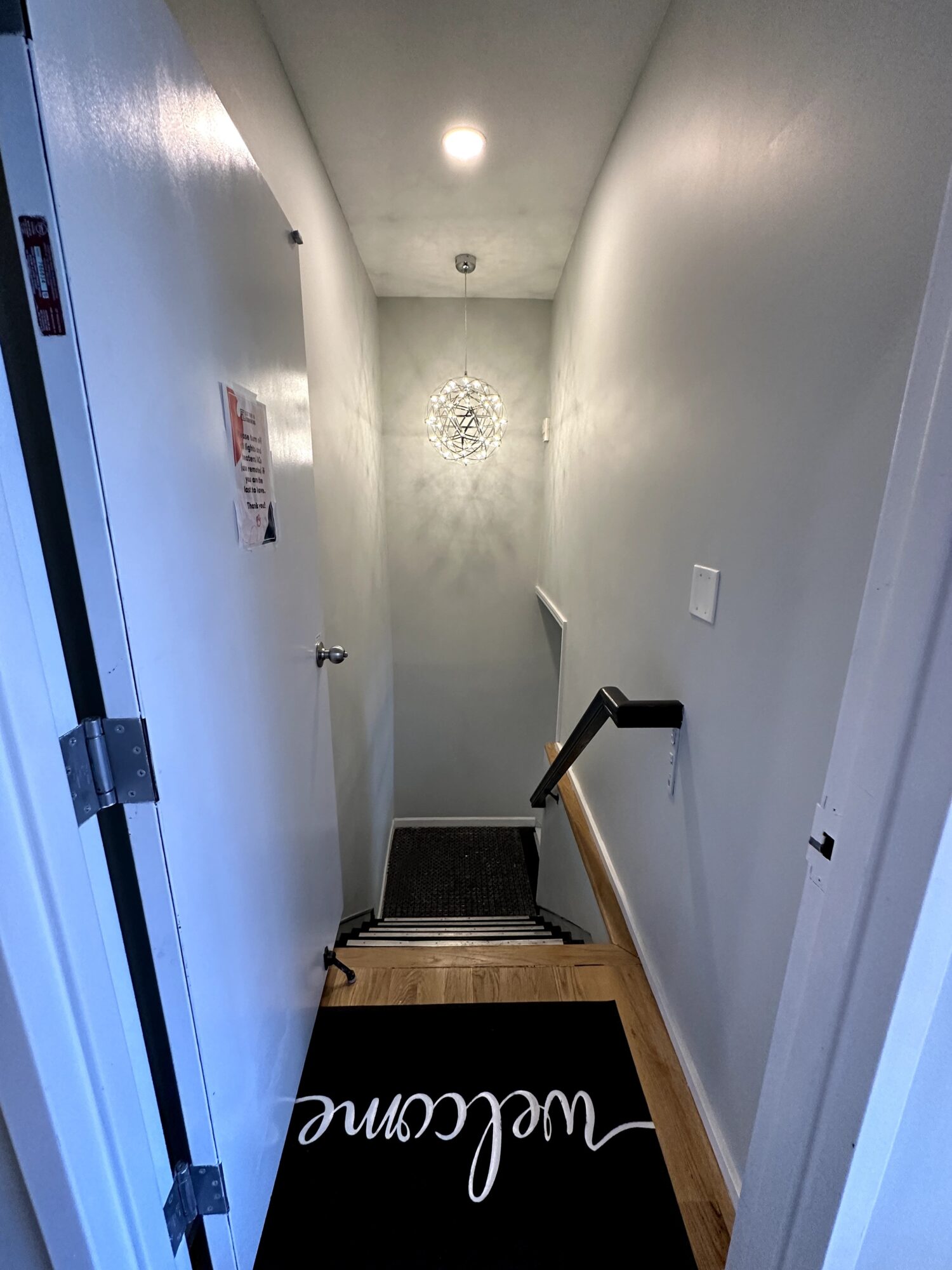 Narrow hallway with white walls, wooden floor, black mat with white text, and a staircase at the end.