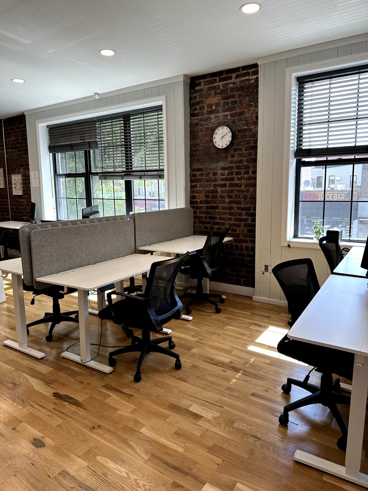 Office space with large windows, a brick wall, a clock, a long table, and chairs, with hardwood floors and natural light.