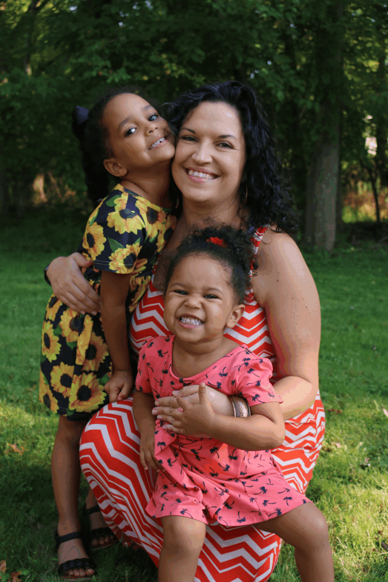 Three people smiling outdoors, two children and an adult woman, with trees and grass in the background.