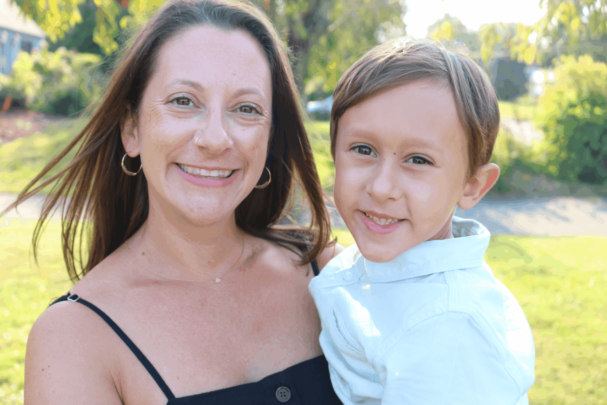 A woman and a young boy smiling outdoors with trees and sunlight in the background.