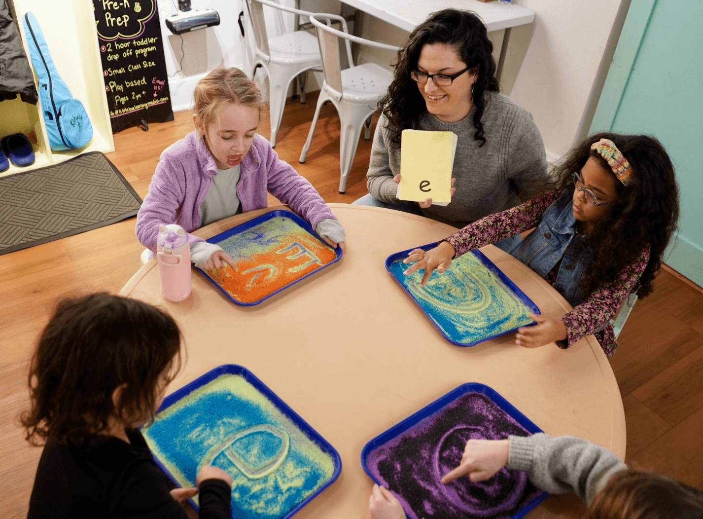 Four children and an adult sit around a table with colorful sand art trays, engaging in a craft activity.