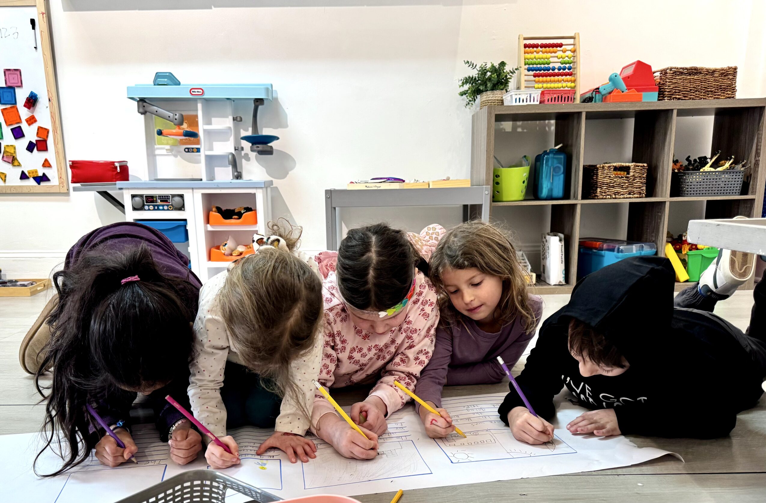 Five children draw on paper at a table in a classroom with shelves and toys in the background.