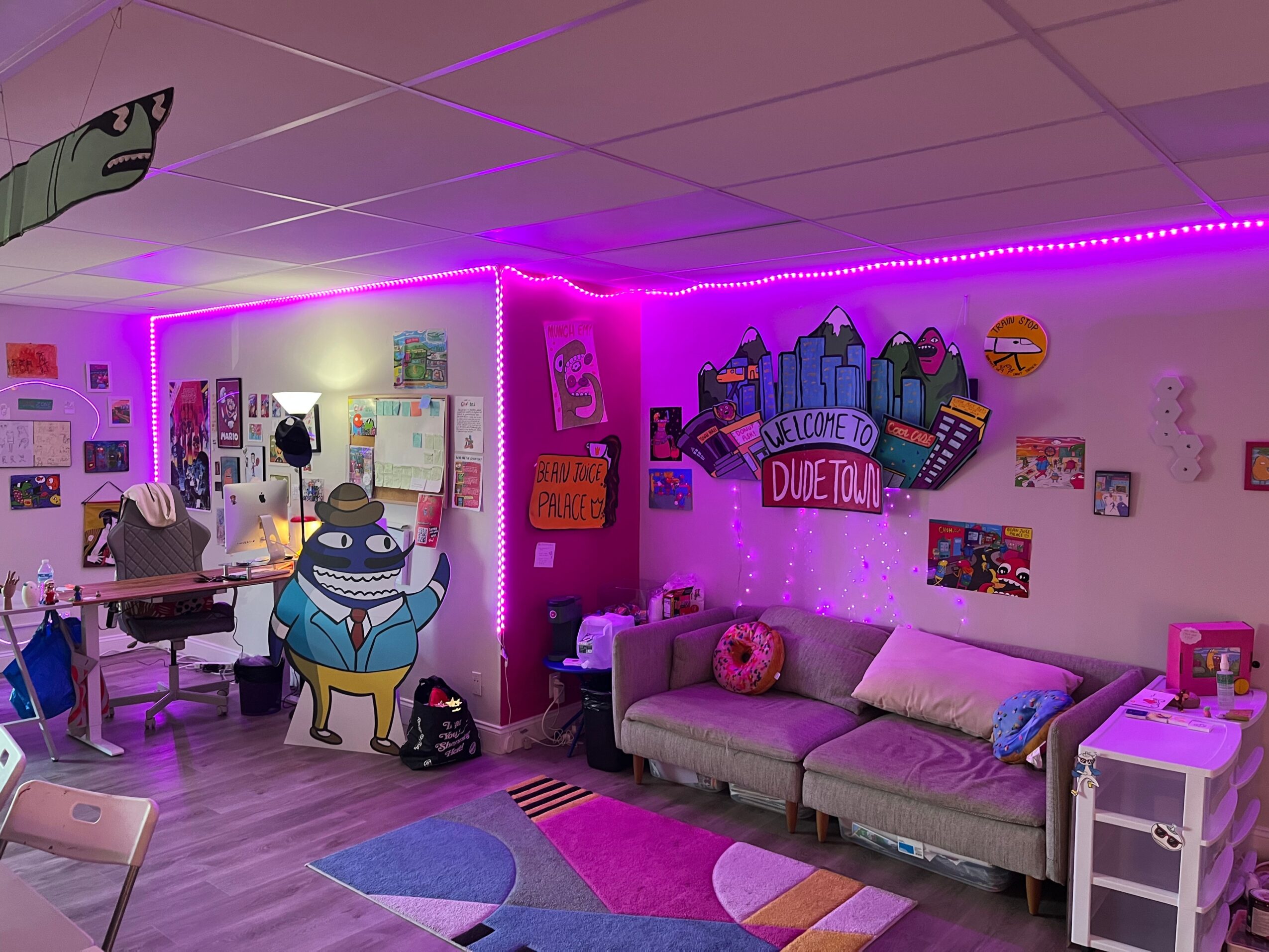 Room decorated with colorful posters, a gray sofa with pillows, a small white table, and neon purple lighting.