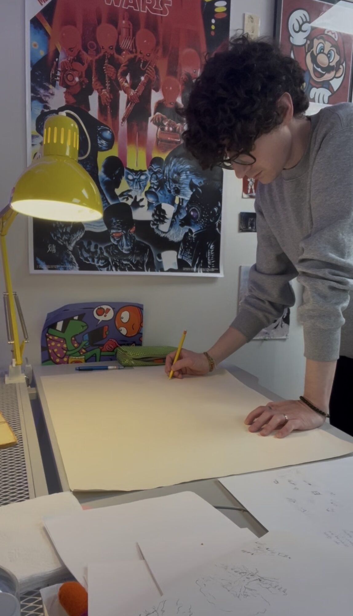 Person with curly hair writing on a large sheet of paper at a desk, posters on the wall behind.
