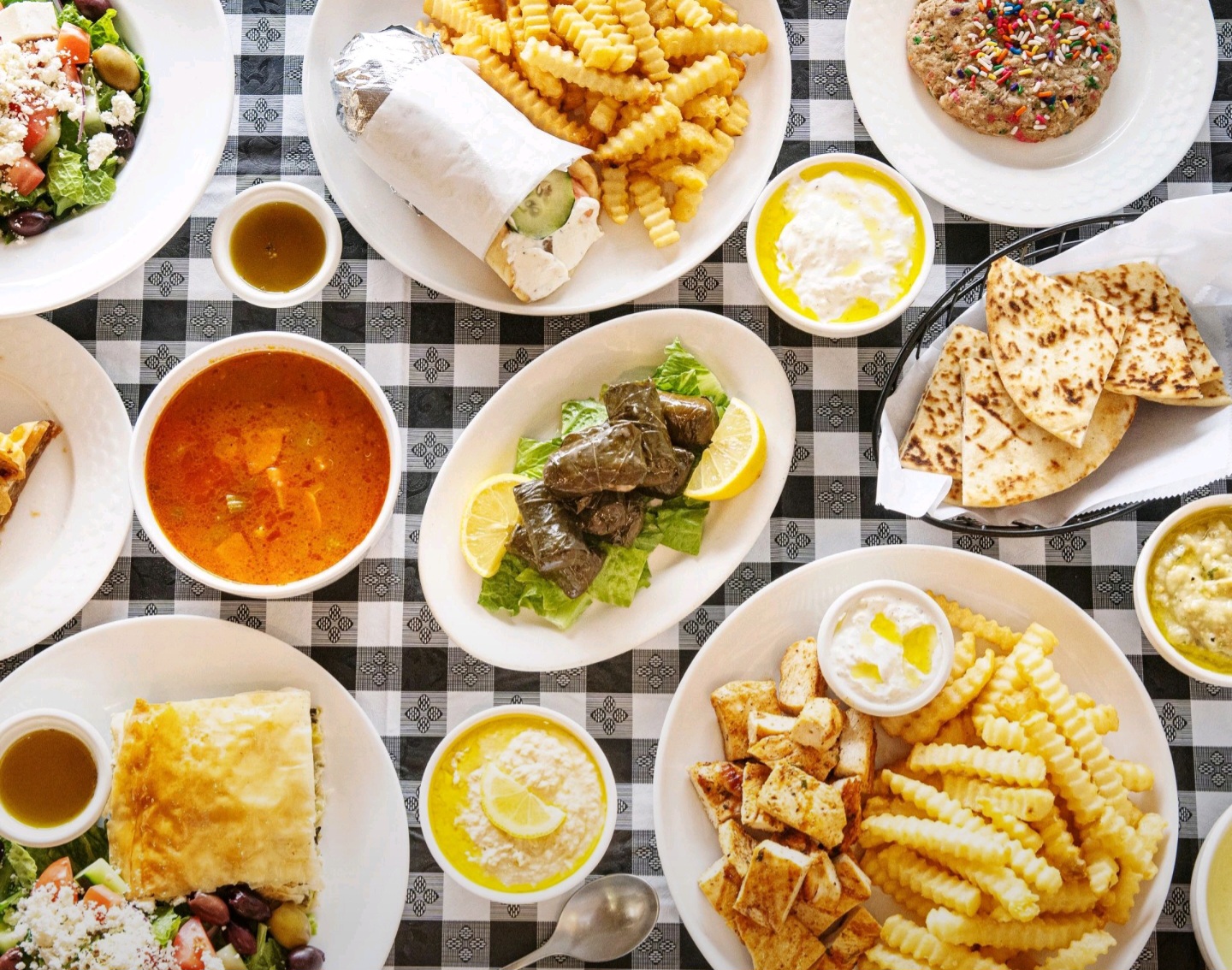 Assorted dishes including fries, salads, bread, and sauces on a checkered tablecloth, viewed from above.