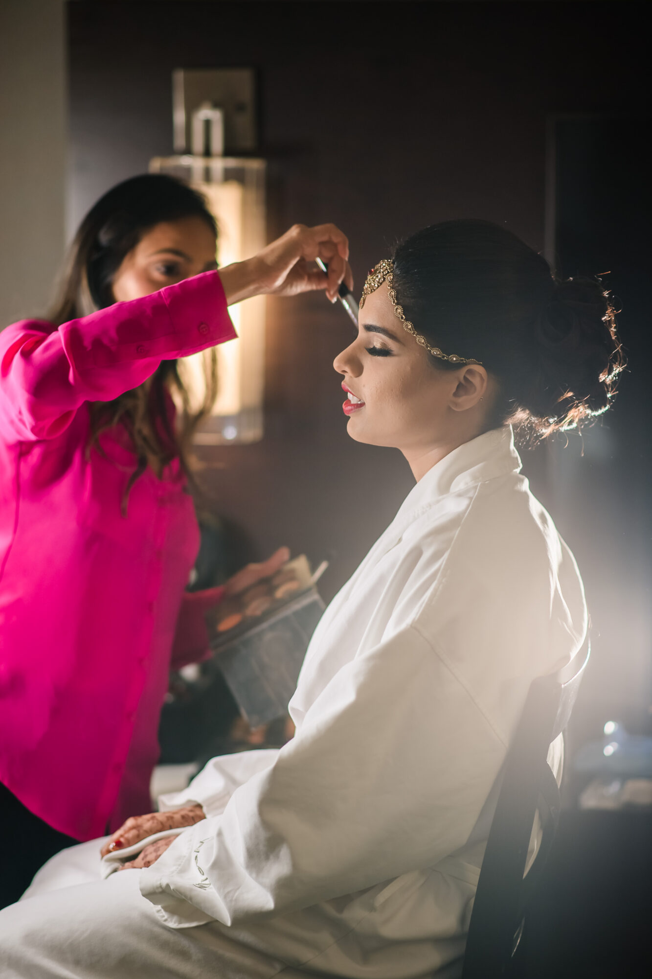 Makeup artist applying makeup to a woman seated in a dimly lit room.
