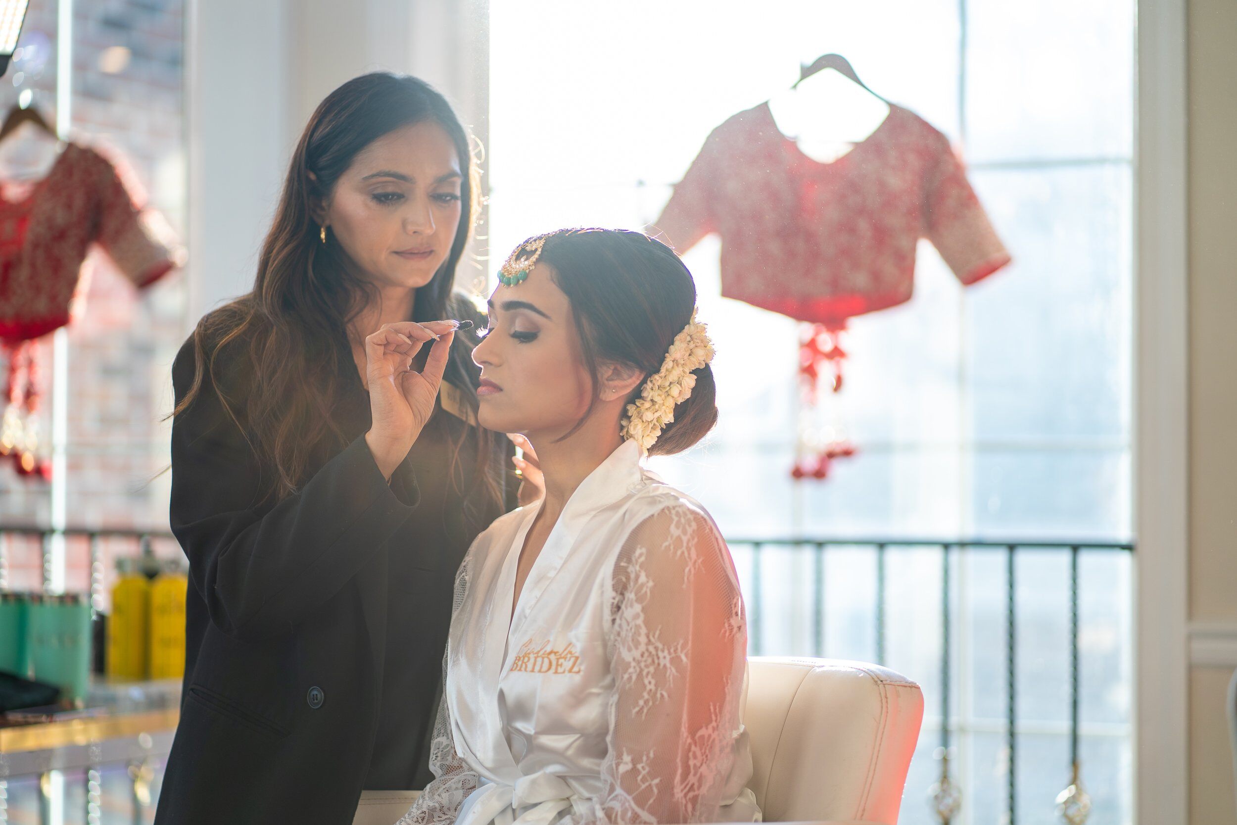 Woman in black applying makeup to a bride in white lace dress with floral hair accessories, in a room with hanging red garments.
