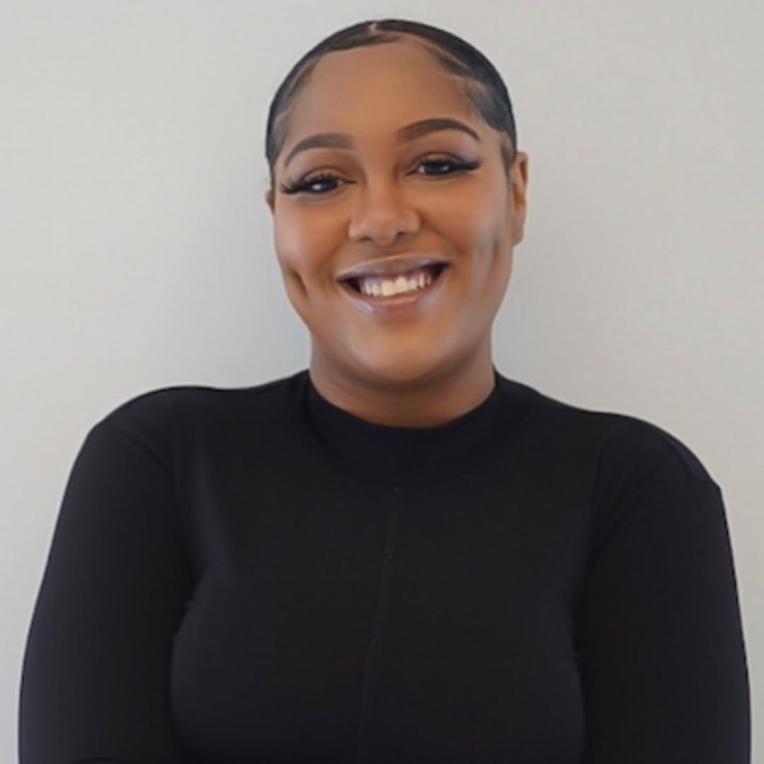 Smiling woman with makeup and slicked-back hair, wearing a black top, against a plain light background.