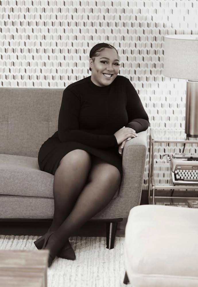 Woman sitting on a sofa, smiling, with a textured wall and lamp in the background.