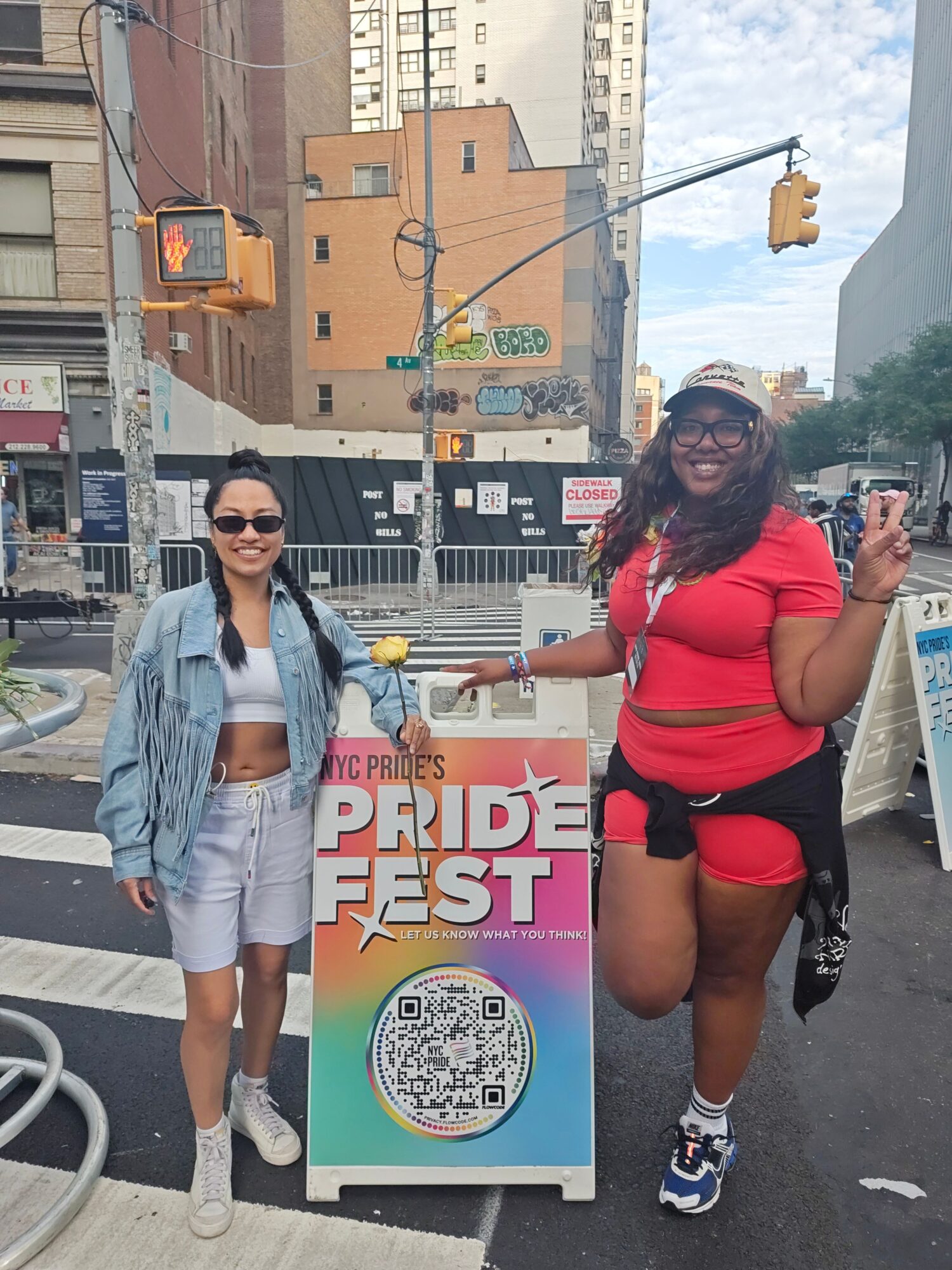 Two women standing on a city street with buildings and traffic lights in the background. One woman wears a red outfit, the other a gray jacket.