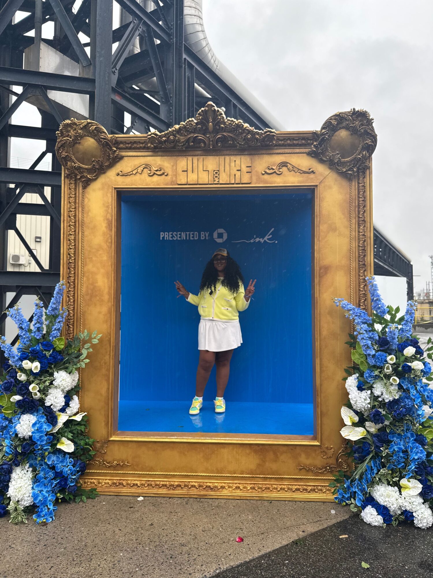Person in a white shirt and shorts standing inside a large ornate gold frame with blue background and flowers at the top and bottom.